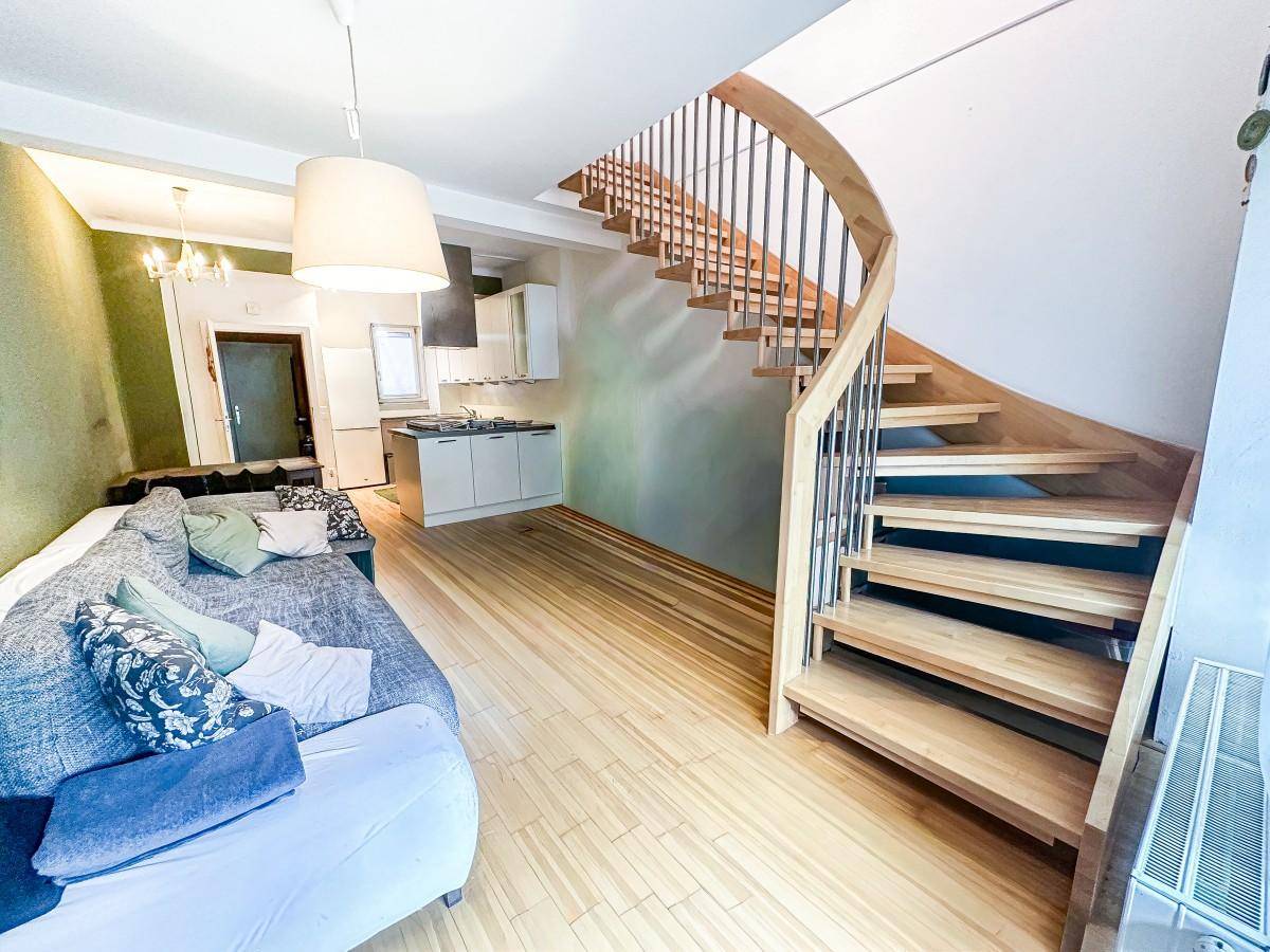 Heller Wohnbereich mit Sofa, Holztreppe und offener Küchenzeile im Hintergrund.
