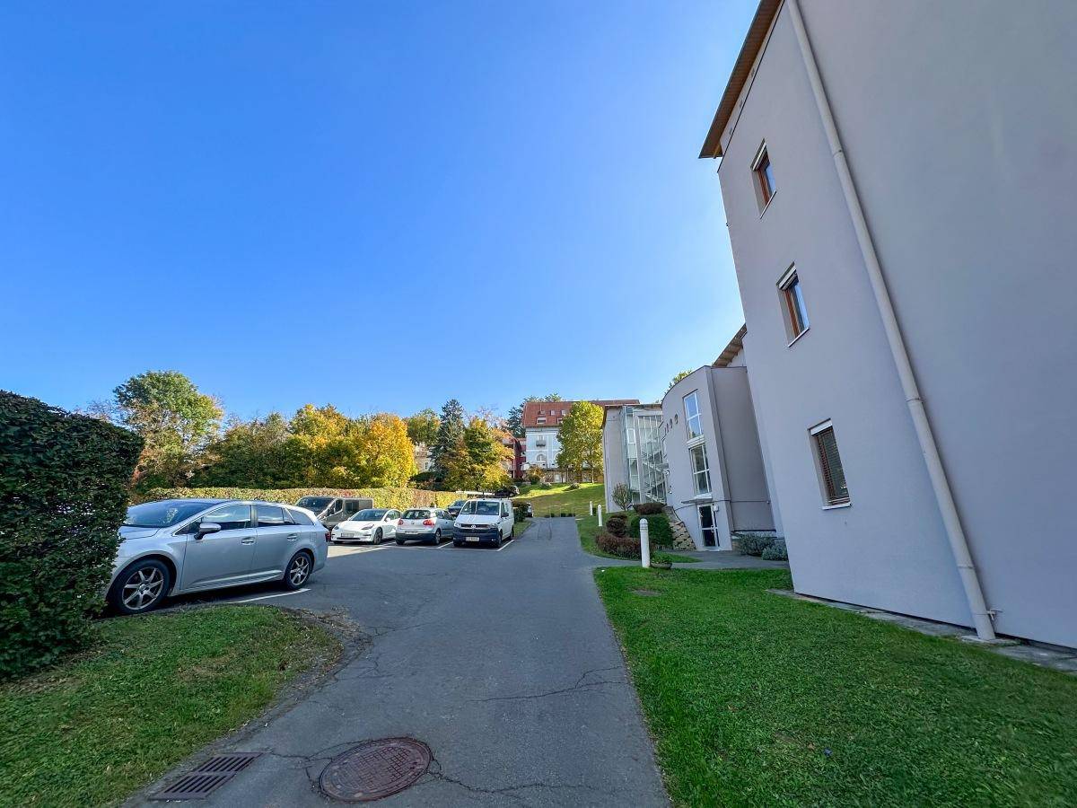 Außenbereich mit Parkplätzen und Zufahrt zum modernen Gebäude, umgeben von Bäumen.