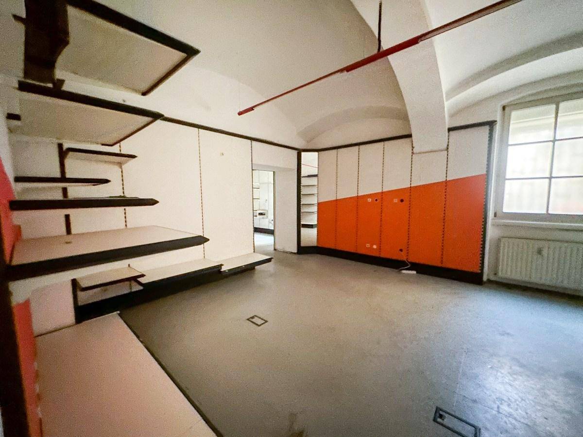 Leerer Ladenraum mit gewölbter Decke, orangefarbenen Akzentwänden und Regalsystemen, mit Fensterblick.