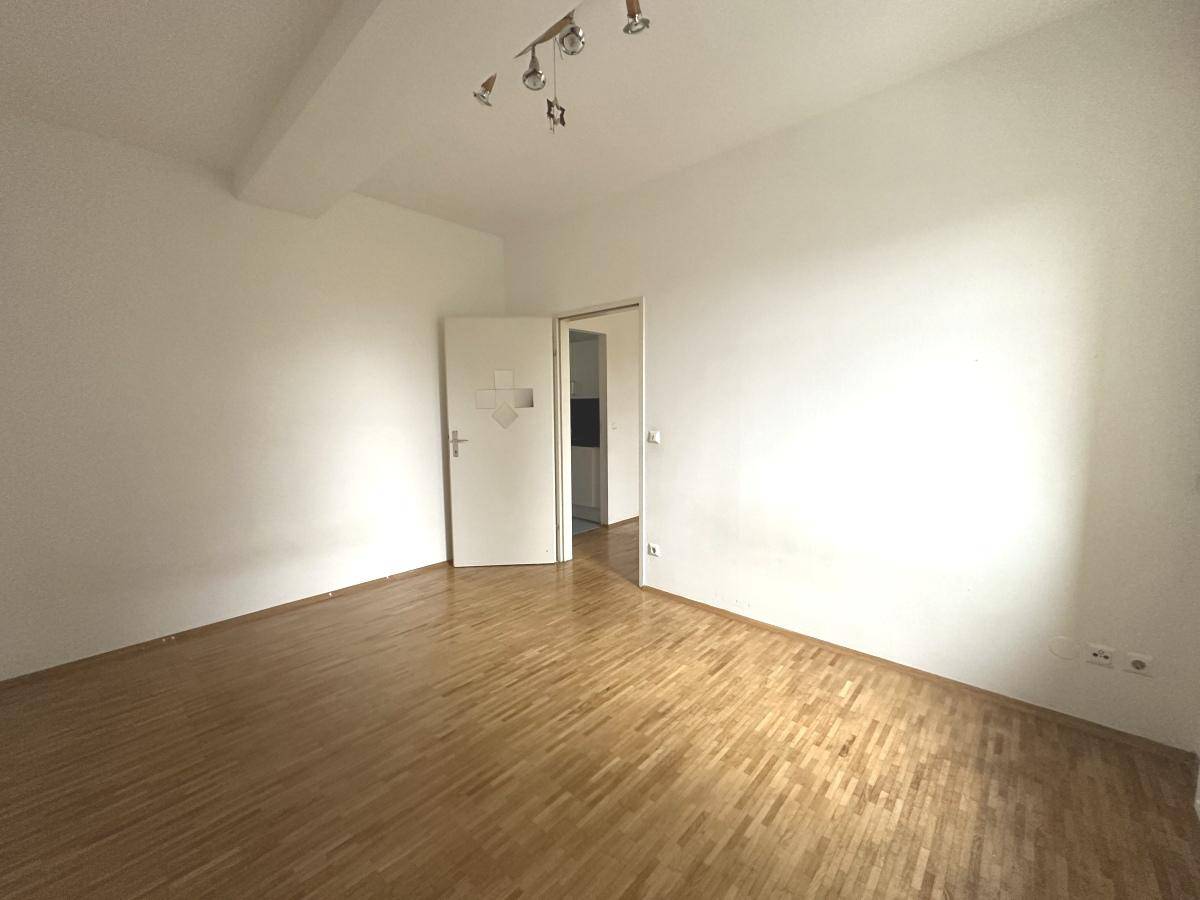 Geräumiges Zimmer mit Parkettboden, weißen Wänden und einer Tür zu einem weiteren Raum.