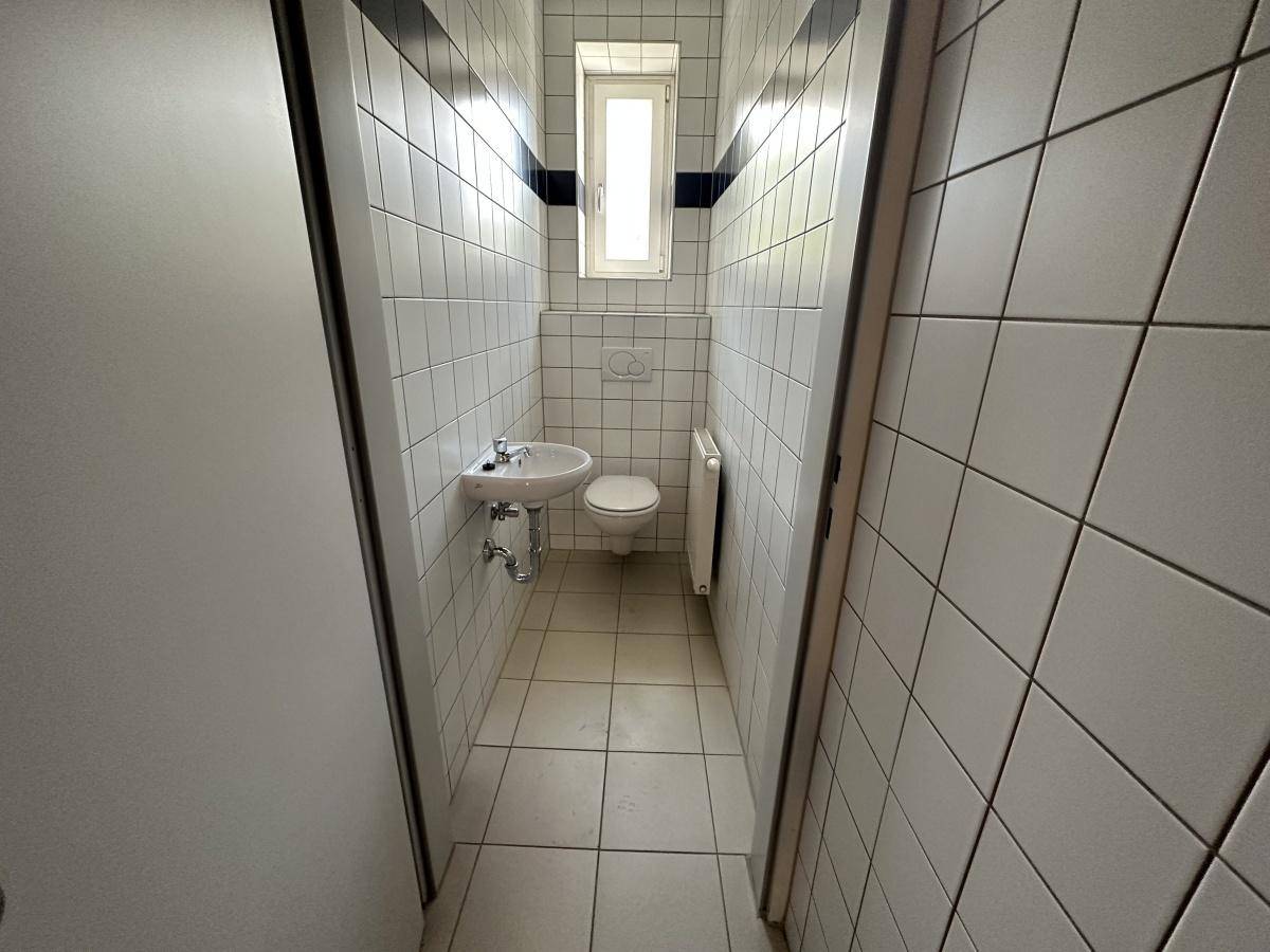 Schmale Toilette mit weißen Fliesen, kleinem Fenster, Waschbecken und WC.