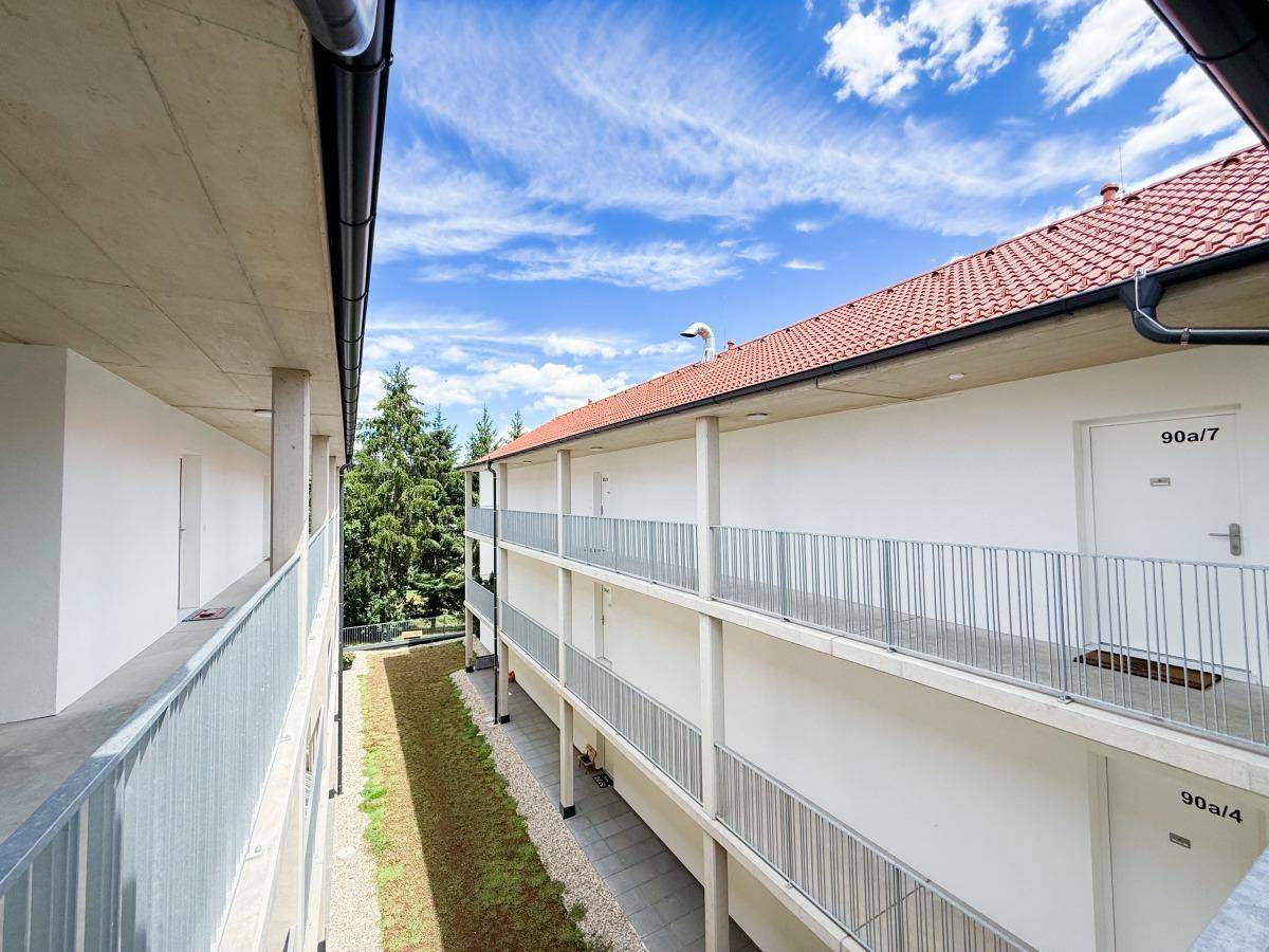 Außenansicht eines Neubaus mit Laubengängen und rotem Ziegeldach unter blauem Himmel.