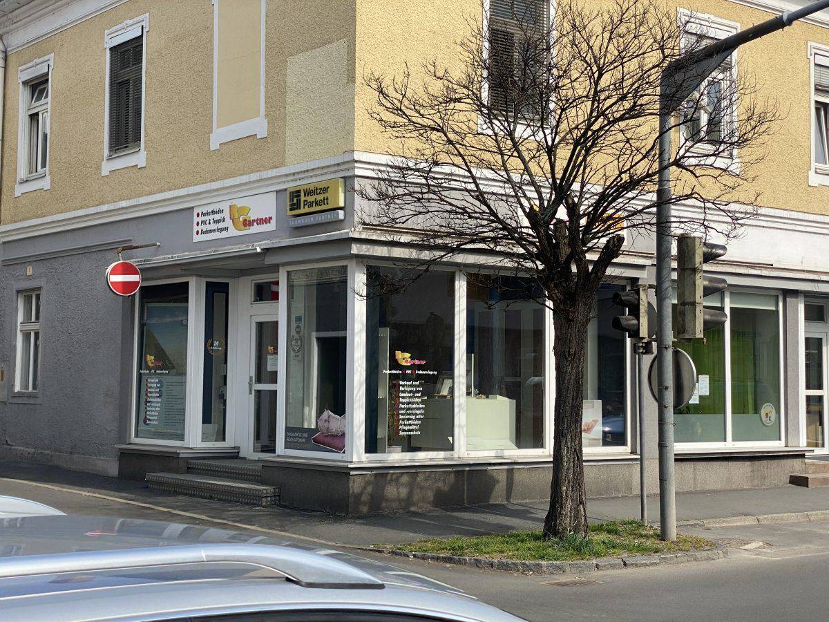 Weitwinkelansicht des Eckgebäudes mit Ladenfront, umgeben von einer Straße und einem kahlen Baum.
