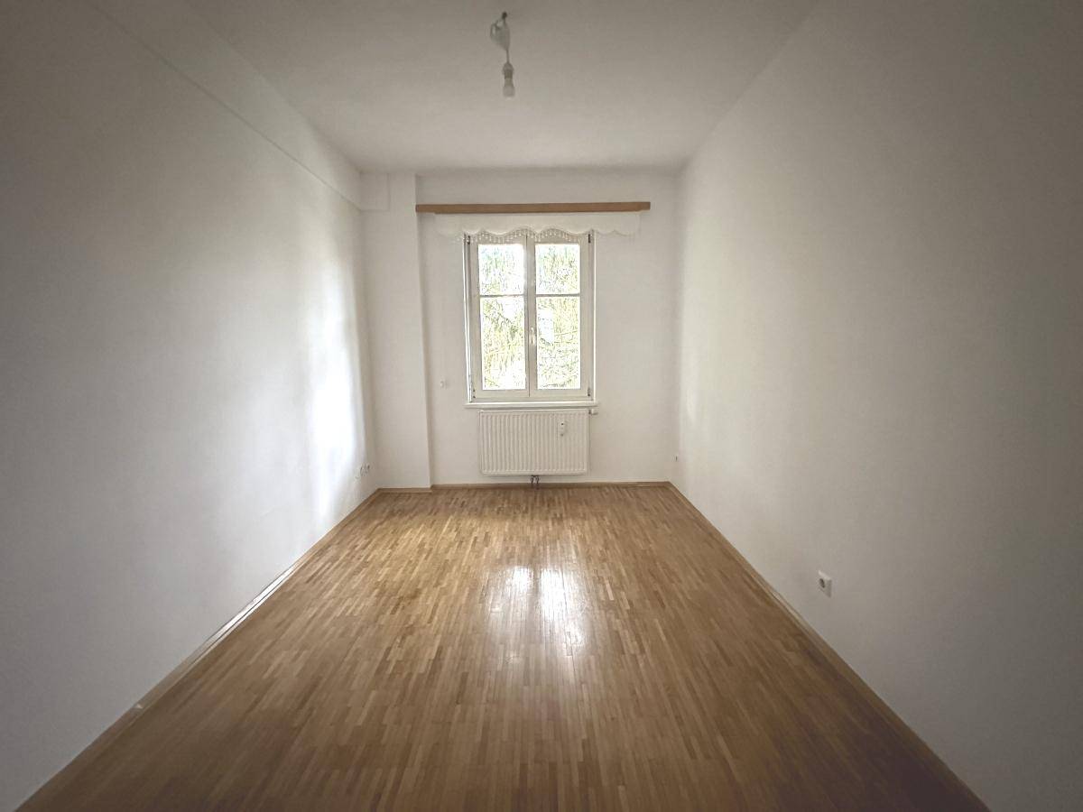 Langes, helles Zimmer mit Parkettboden und einem Fenster, das viel Licht spendet.