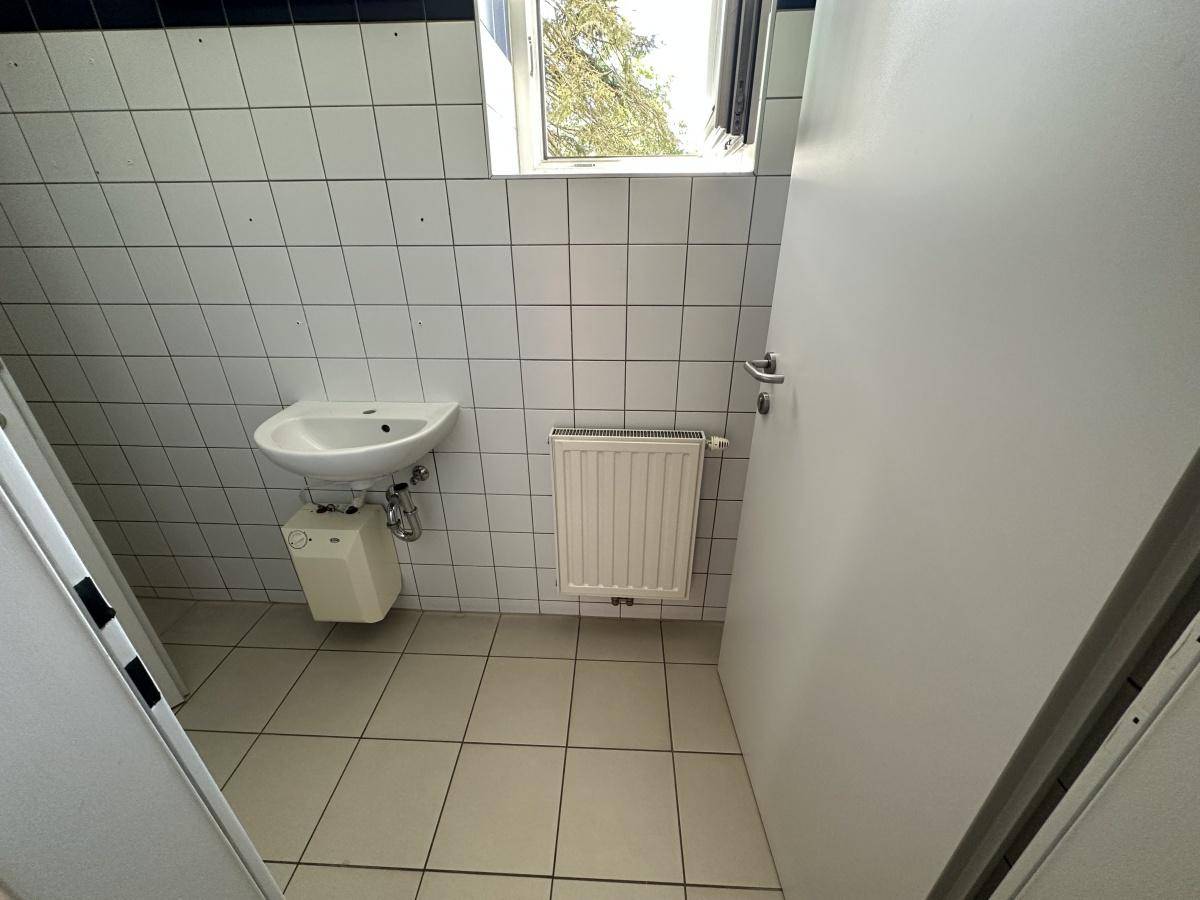 Toilette mit weißen Fliesen, Fenster, Waschbecken und Heizkörper.