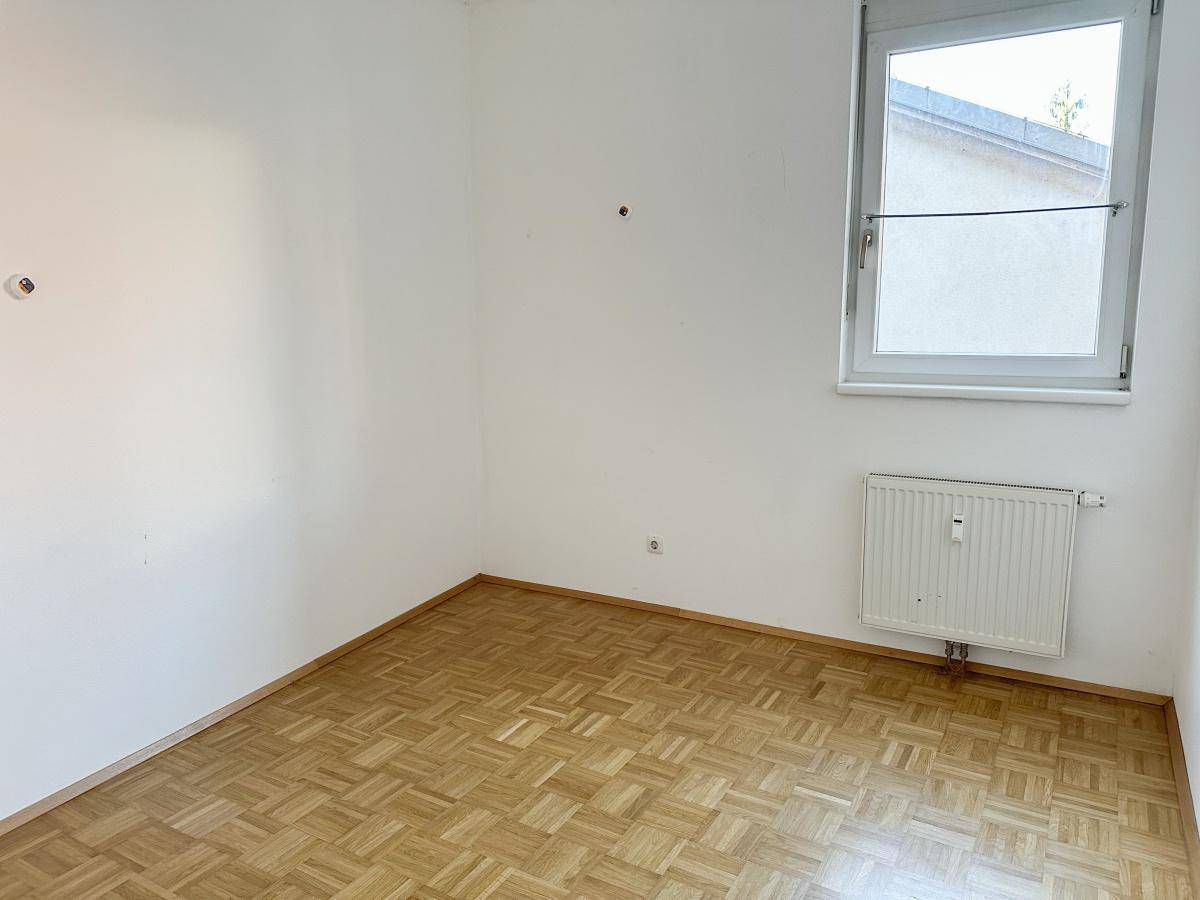 Ruhiges Schlafzimmer mit Parkettboden, weißen Wänden und einem Fenster mit Blick nach draußen.