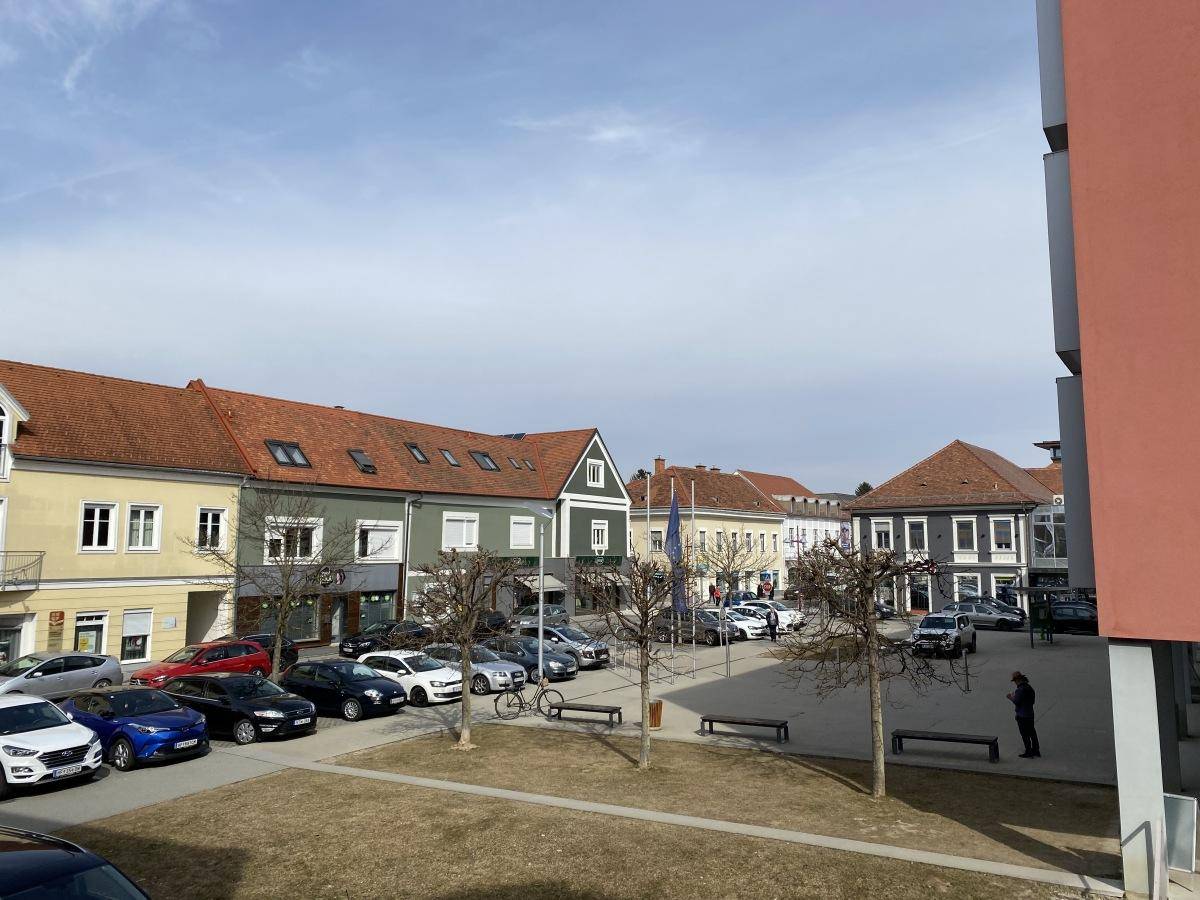 Blick auf einen belebten Stadtplatz mit parkenden Autos und traditionellen Gebäuden unter einem klaren blauen Himmel.