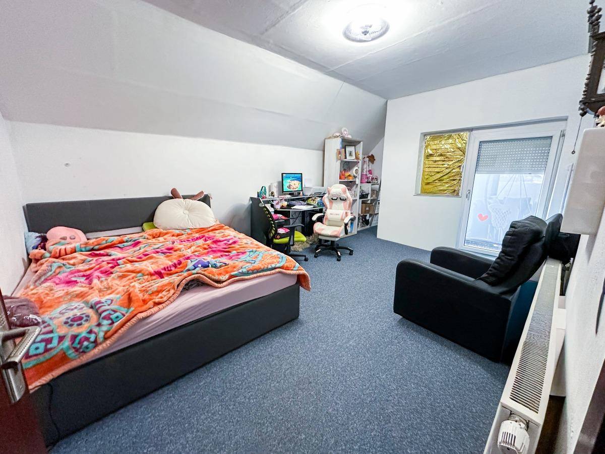 Schlafzimmer mit blauem Teppichboden, Dachschräge und einem Fenster mit Rollladen.