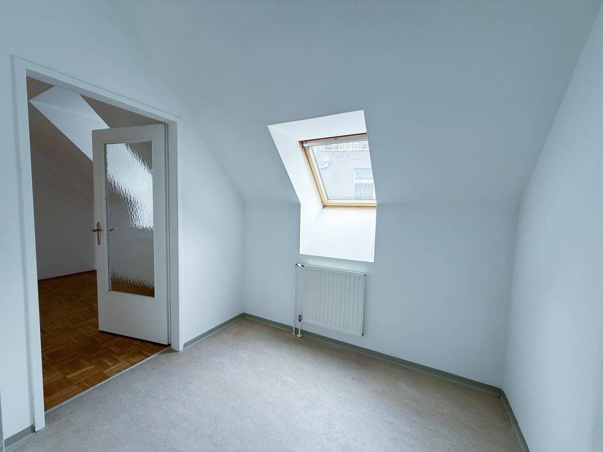 Ruhiges Zimmer mit hellem Bodenbelag, weißen Wänden und einem Dachfenster, ideal als Schlafzimmer.