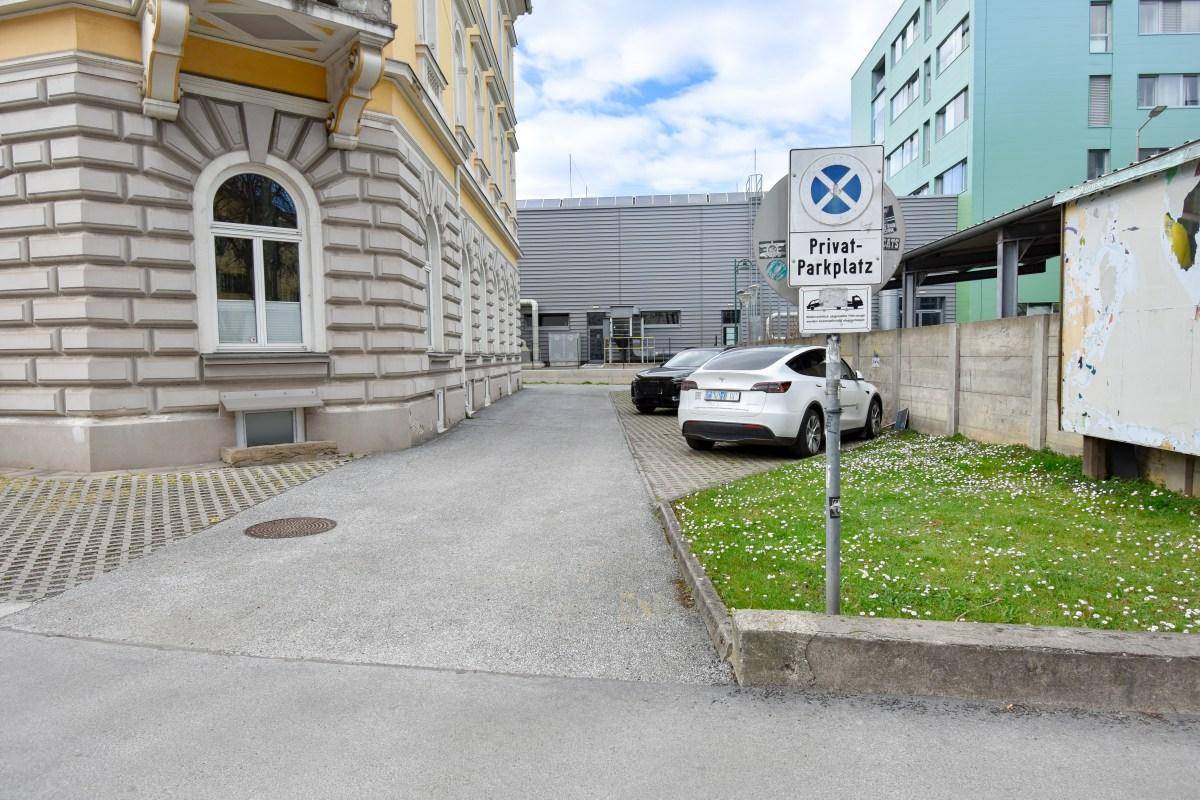 Seitenansicht eines Gebäudes mit einem privaten Parkplatz und angrenzender Grünfläche unter bewölktem Himmel.