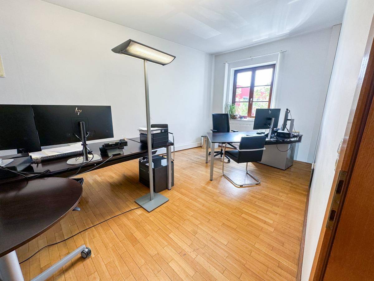 Helles Einzelbüro mit Holzfußboden, großem Fenster und mehreren Computermonitoren.