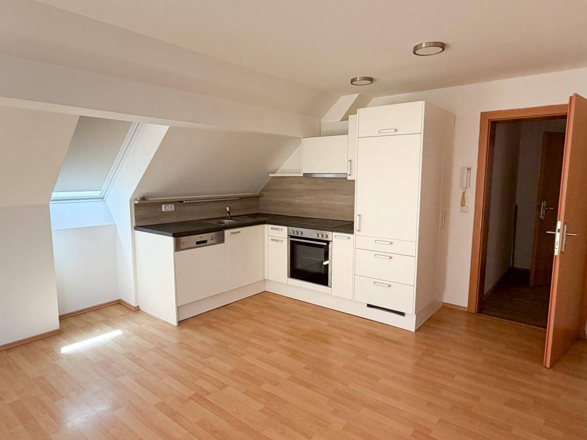 Funktionale Einbauküche in einem Dachgeschossraum mit weißen Schränken, dunkler Arbeitsplatte und Zugangstür.