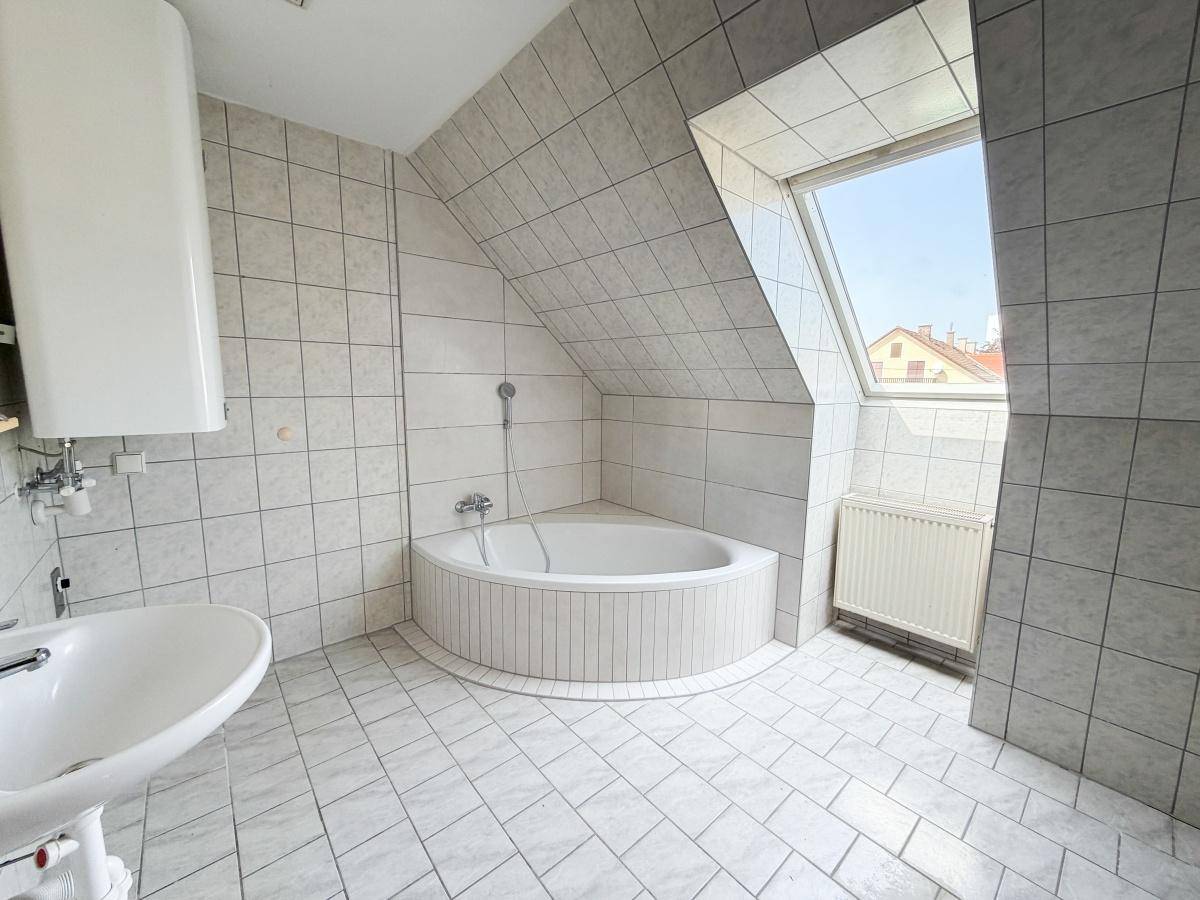 Gefliestes Badezimmer im Dachgeschoss mit Eckbadewanne, Waschbecken, Warmwasserboiler und Dachfenster.