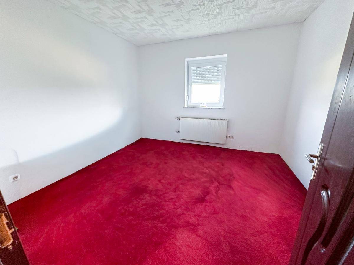 Leeres Schlafzimmer mit leuchtend rotem Teppichboden und Fenster mit Rollladen.