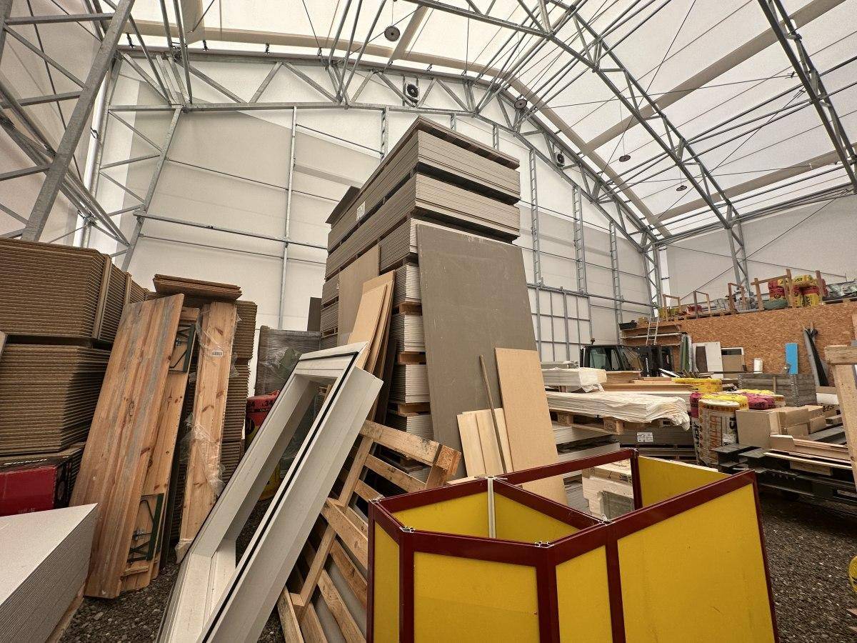 Große Lagerhalle mit Metallkonstruktion und gestapelten Baumaterialien wie Holzplatten und Fensterrahmen.