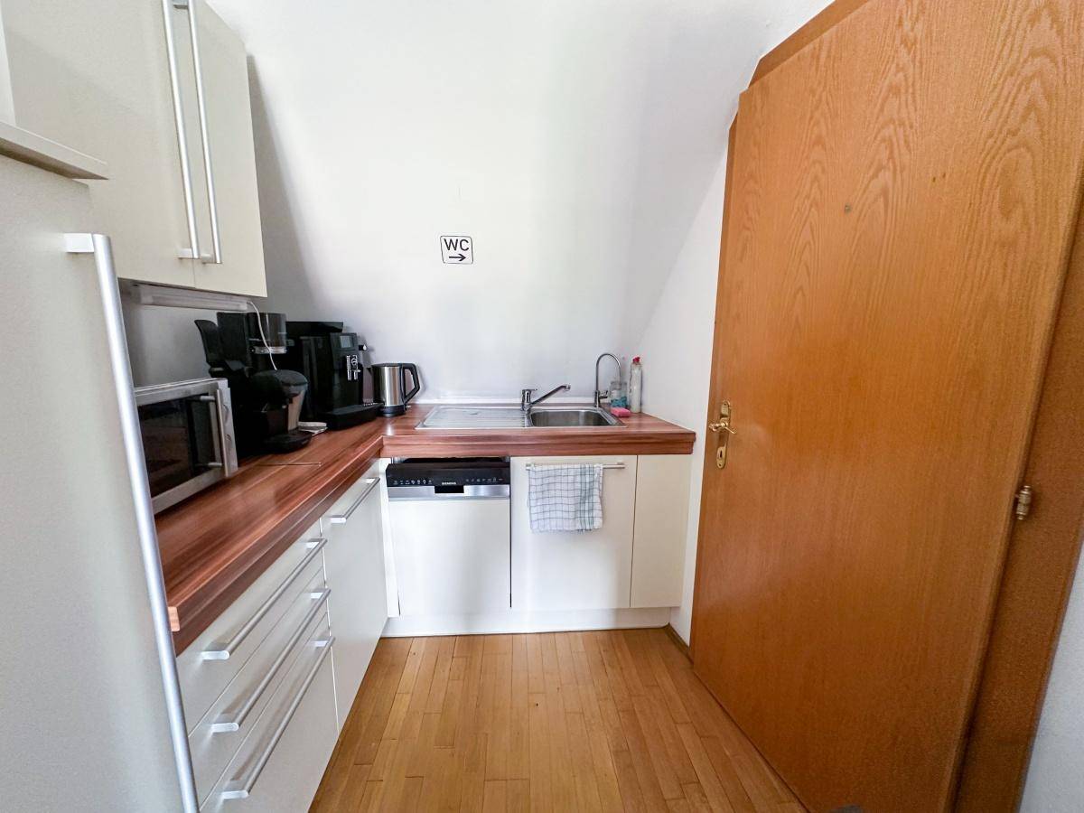 Kleine Teeküche mit Holzarbeitsplatte, Spüle, Kühlschrank und Kaffeemaschine unter Dachschräge.