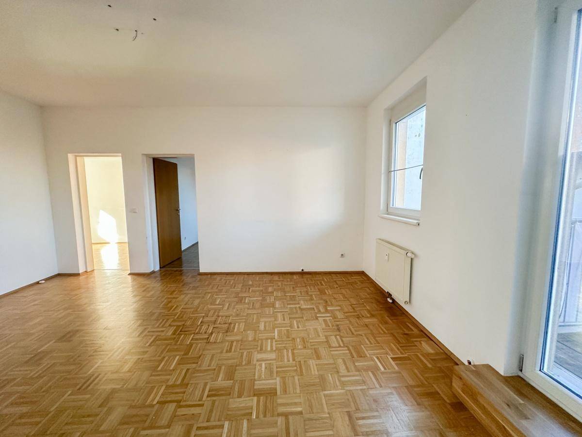 Helles Wohnzimmer mit Parkettboden, weißen Wänden und Zugang zu einem Balkon.