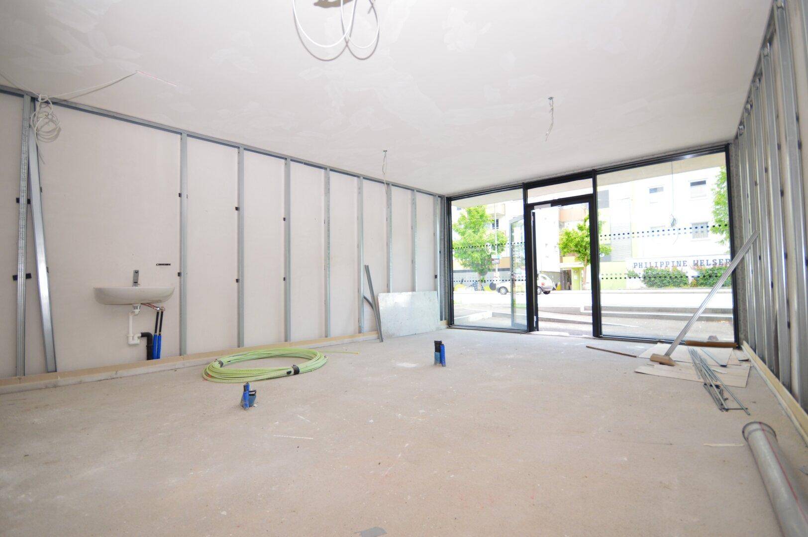 Rohbau eines Raumes mit großen Fenstern, Metallständern und einer vorbereiteten Installation für ein Waschbecken an der Wand.