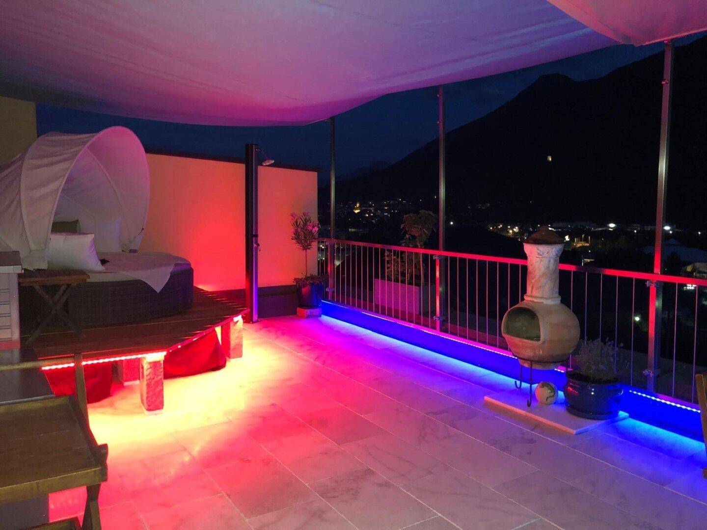 Beleuchtete Terrasse bei Nacht mit Liegemöbeln, Feuerstelle und Blick auf die Stadtlichter.