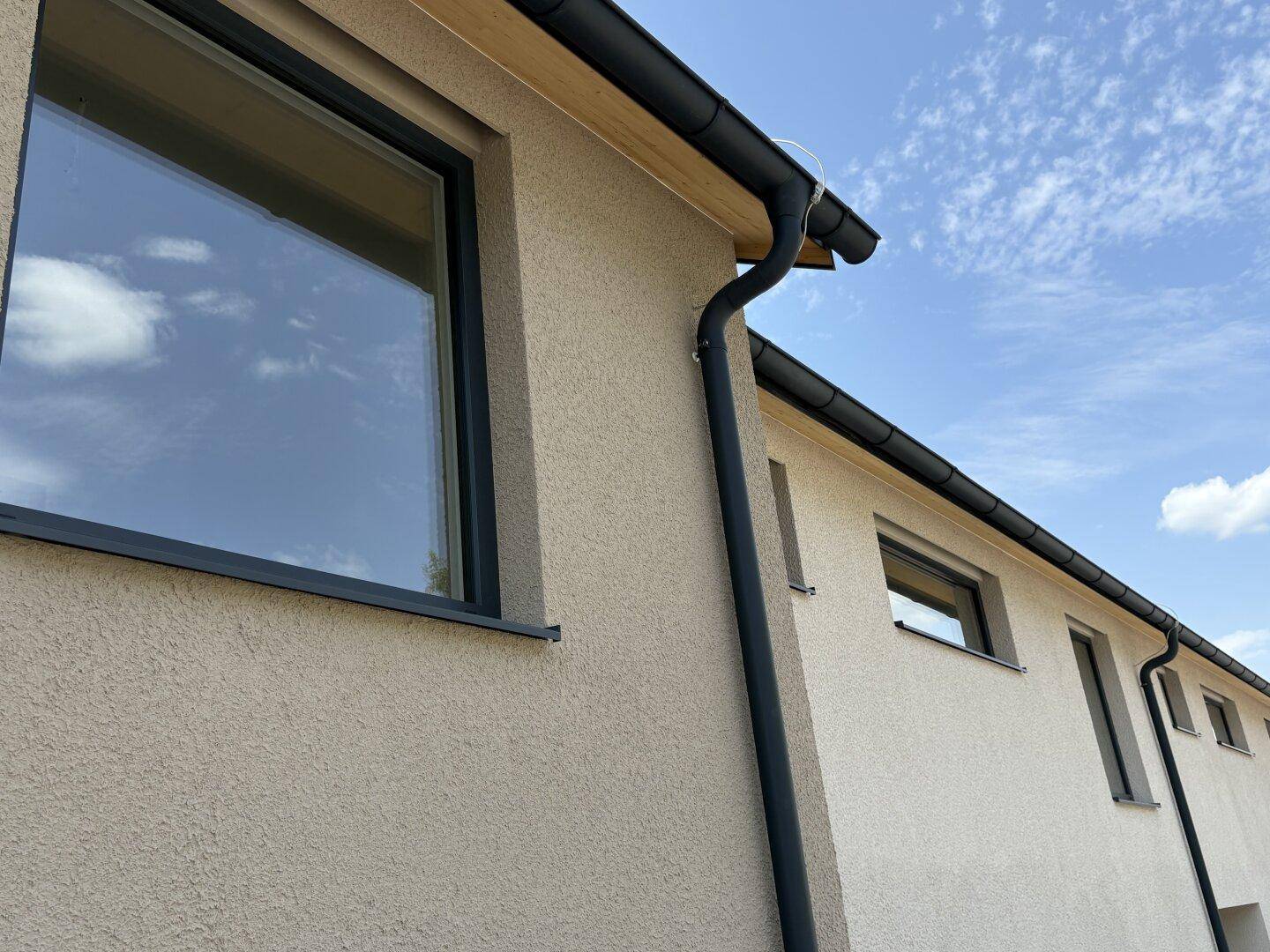 Detailansicht der modernen Fassade mit Putzstruktur, dunklen Fenstern und Regenrinnen unter blauem Himmel.