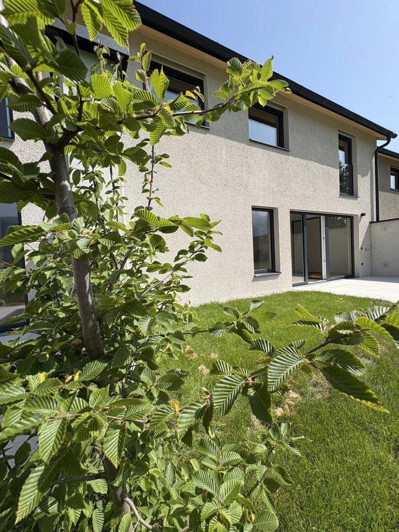 Moderne Hausfassade mit großen Fenstern und gepflegtem Rasen in sonniger Umgebung.