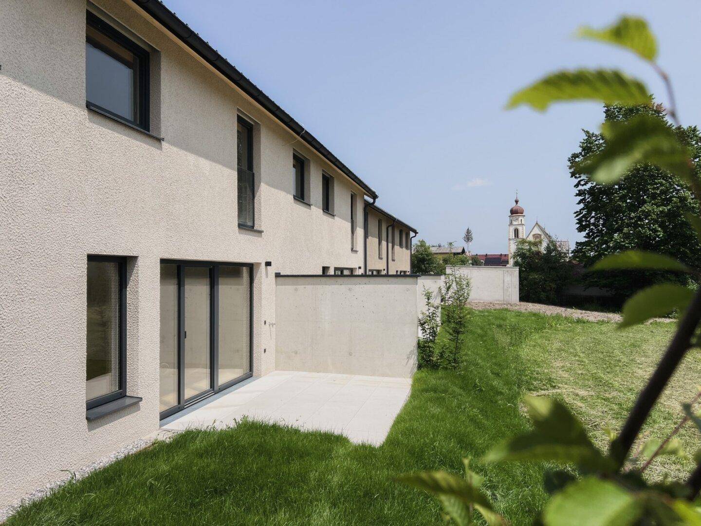 Moderne Gebäudefassade mit großen Fenstern, angrenzender Rasenfläche und Blick auf eine Kirche.
