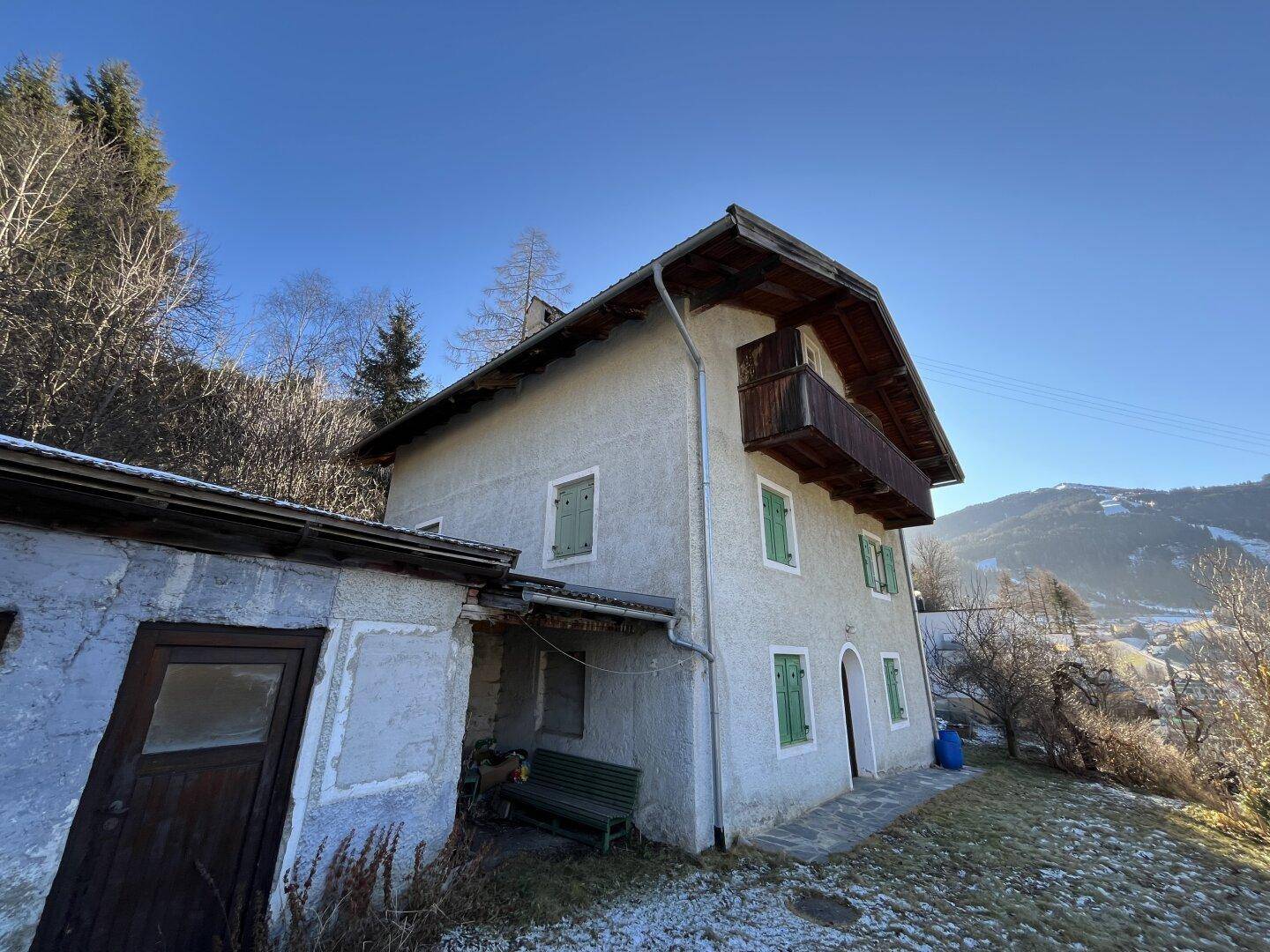 Seitenansicht des traditionellen Hauses mit Anbau und Balkon in sonniger Berglandschaft.