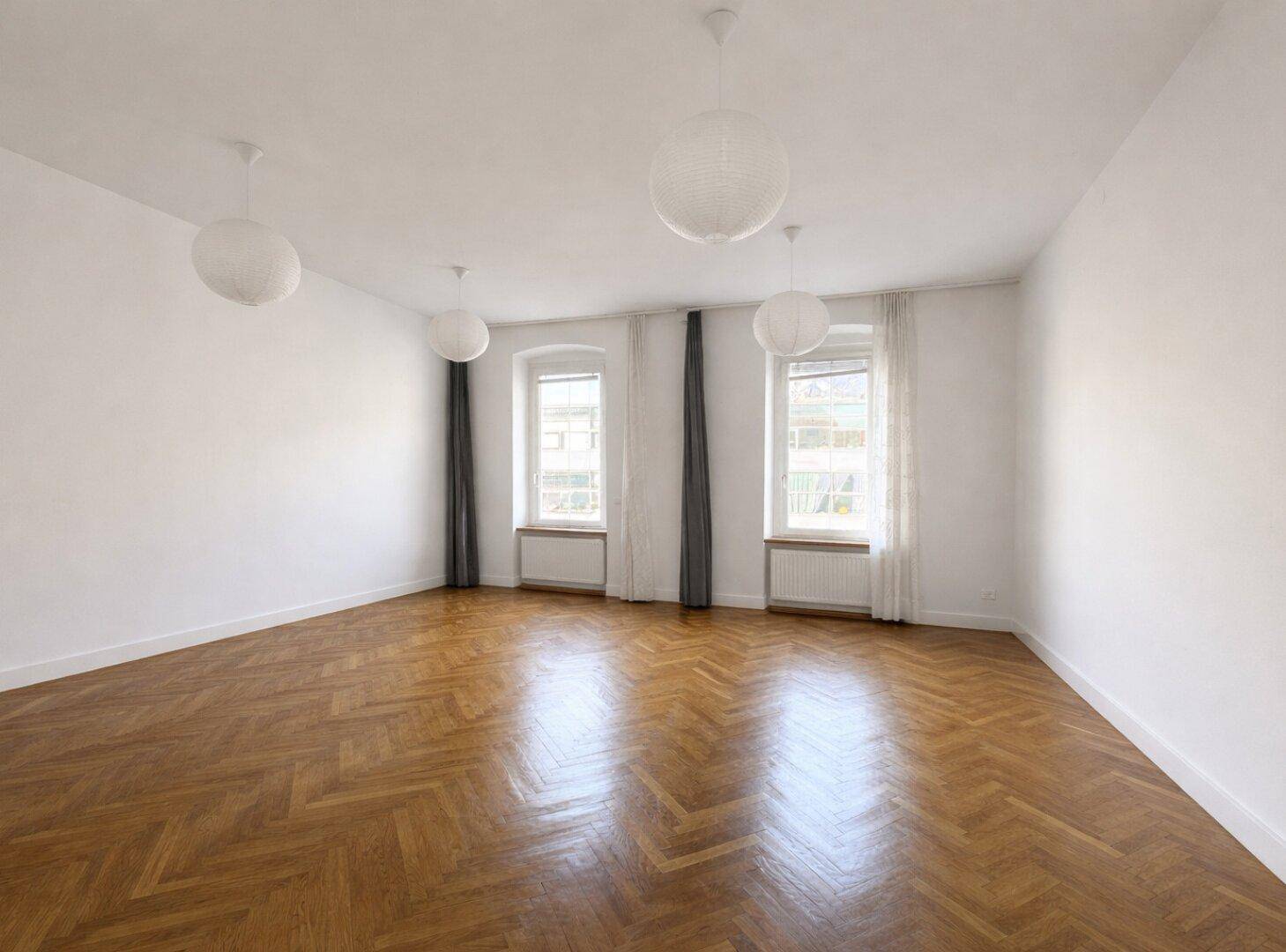 Helles Wohnzimmer mit Fischgrätparkett, hohen Decken und zwei großen Fenstern.