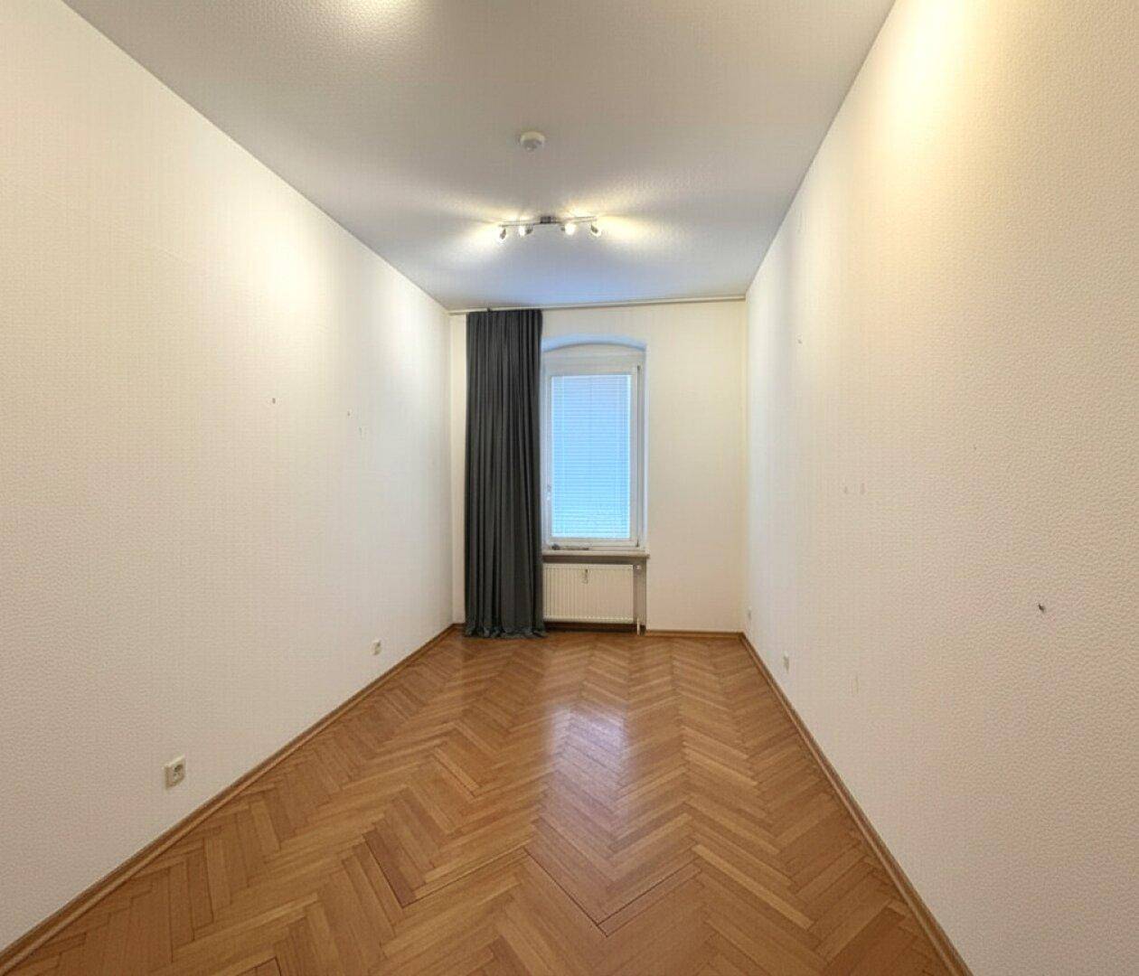 Schmales Zimmer mit Fischgrätparkett, weiß gestrichenen Wänden und einem Fenster.