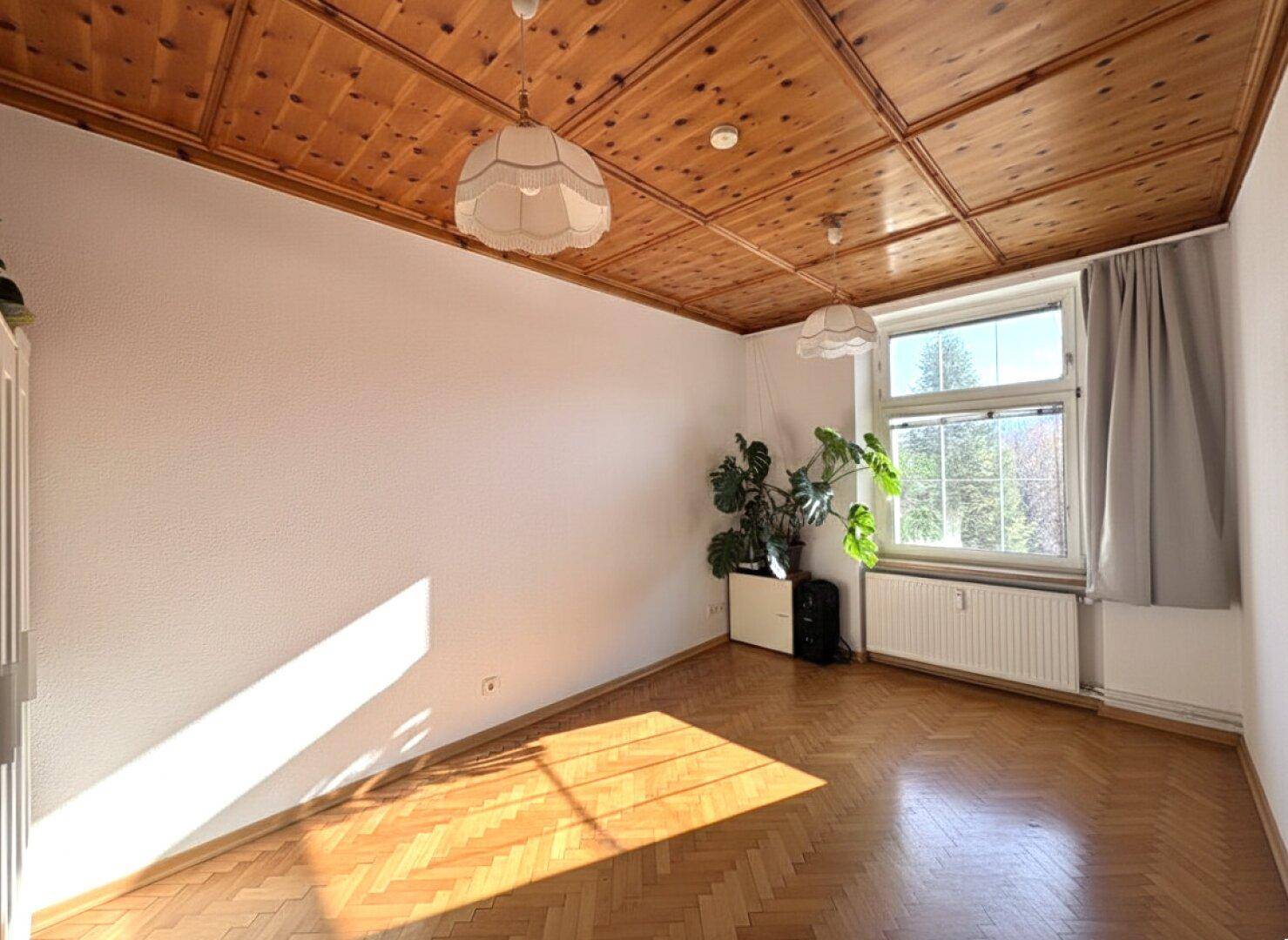 Helles Zimmer mit Holzdecke, Fischgrätparkett und großem Fenster mit Grünblick.
