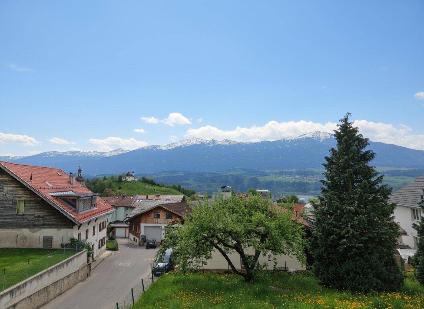 Weitblick über die grüne Landschaft mit Häusern und majestätischen Bergen im Hintergrund unter blauem Himmel.