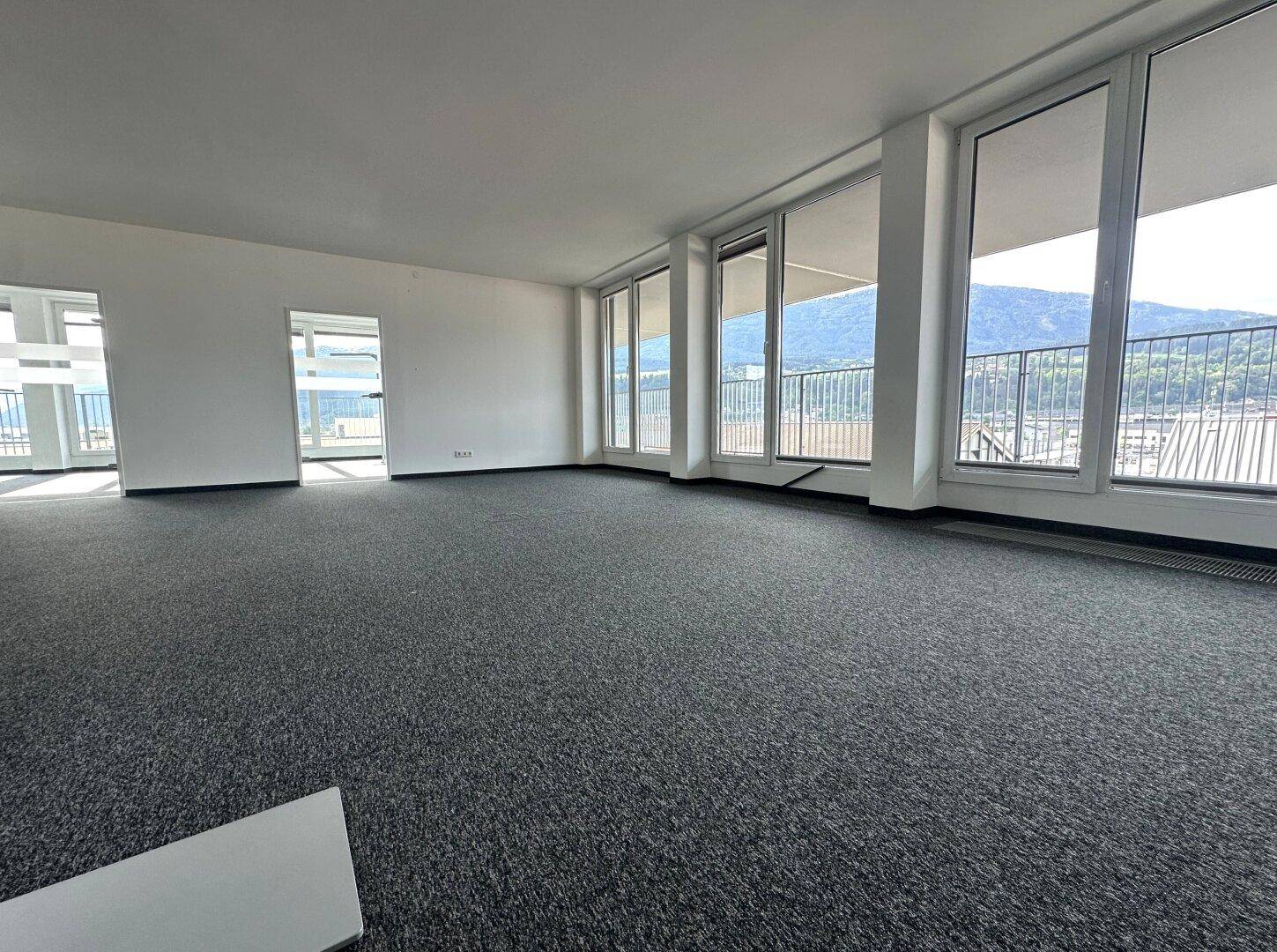 Großer, leerer Büroraum mit Teppichboden und weitem Blick auf die Berge durch die Fensterfront.