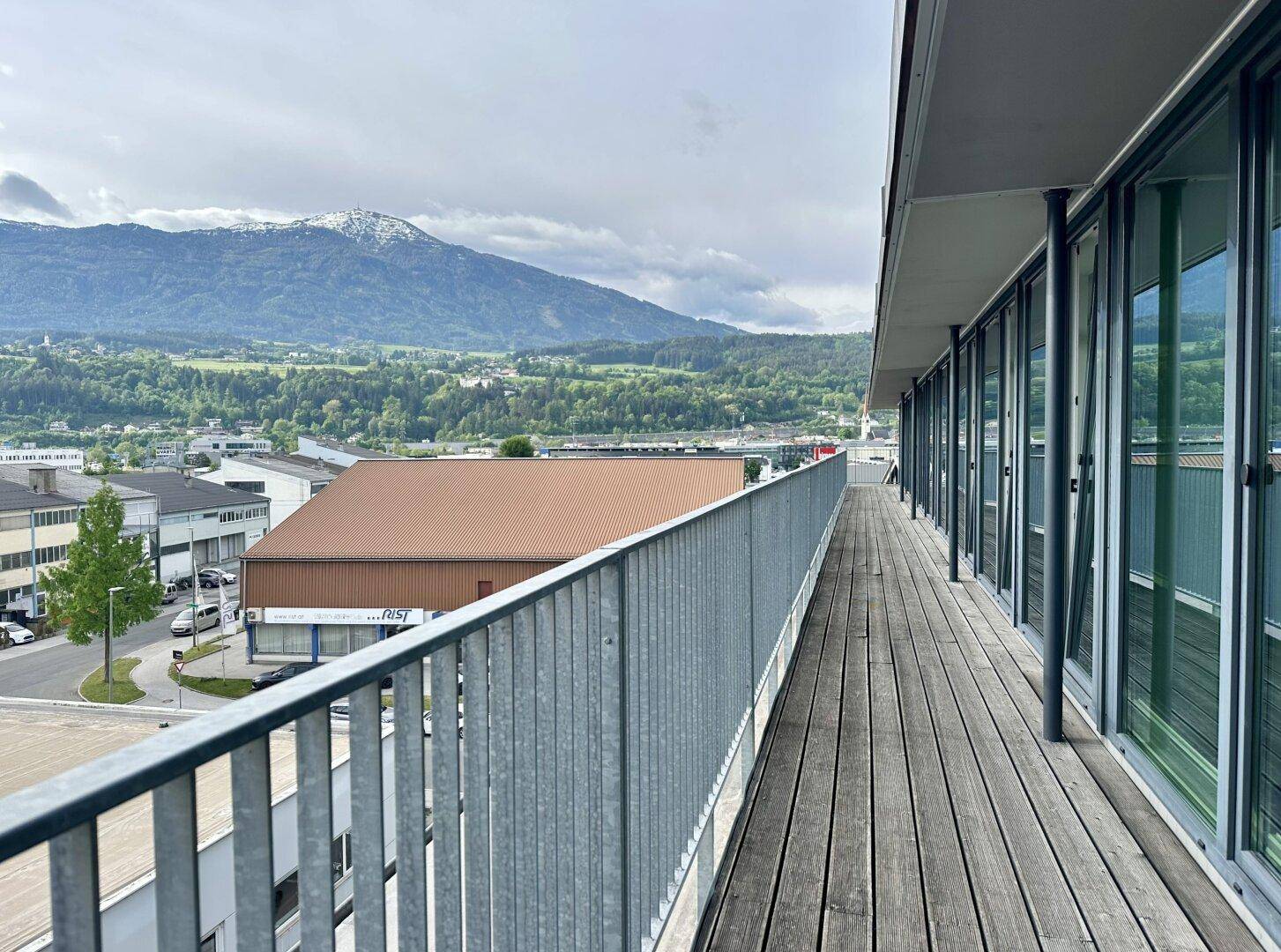 Lange Holzterrasse mit Metallgeländer und weitem Blick über die Stadt und die umliegenden Berge.