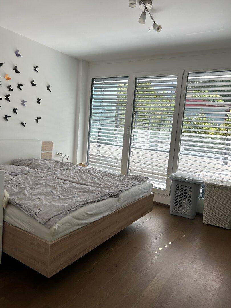 Helles Schlafzimmer mit Doppelbett, Holzboden und großen Fenstern mit Jalousien.