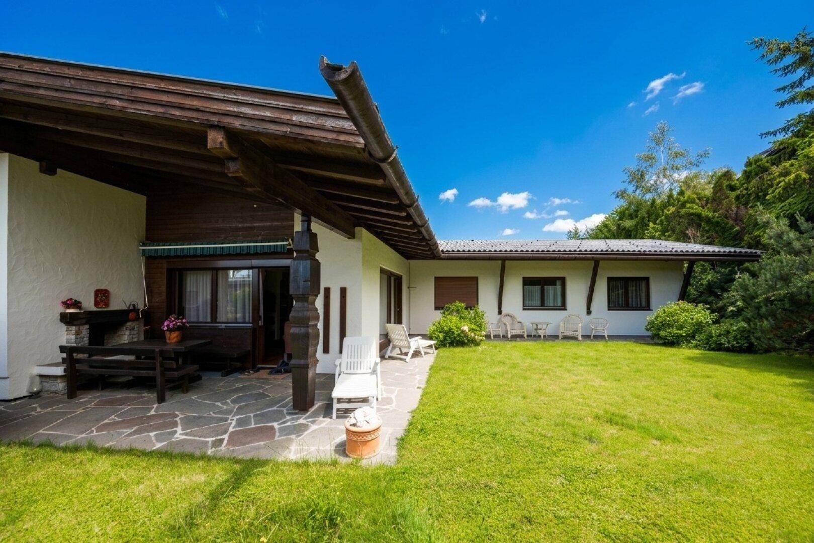 Große überdachte Terrasse mit Steinfliesen und rustikalen Holzmöbeln im Gartenbereich.