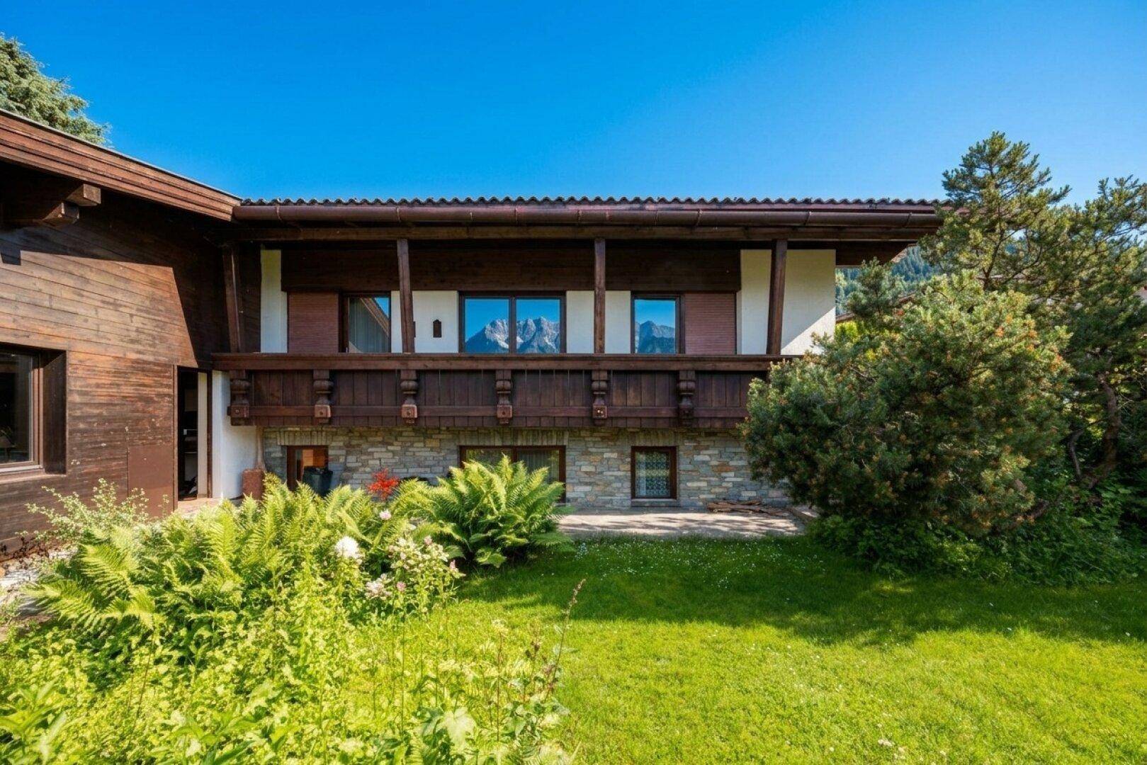 Traditionelle Holzfassade mit Balkon und Steinelementen, umgeben von üppigem Grün und Pflanzen.