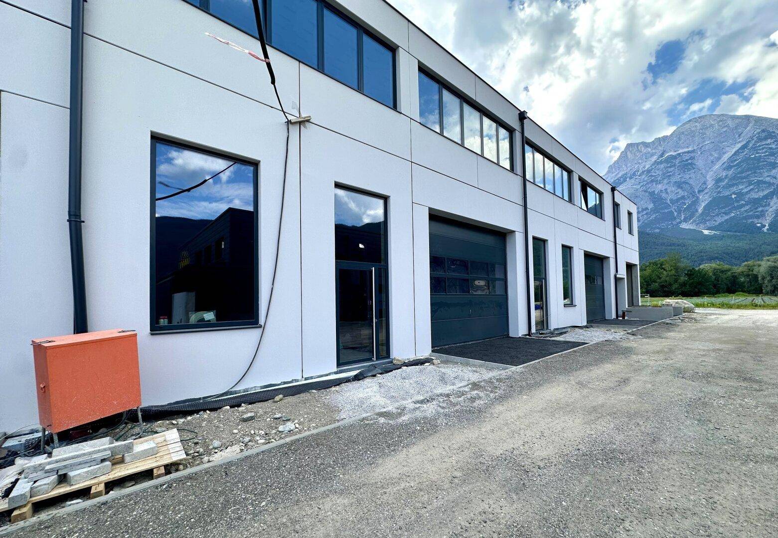 Seitenansicht des Neubaus mit mehreren Garagentoren und Fenstern, umgeben von Bergen.
