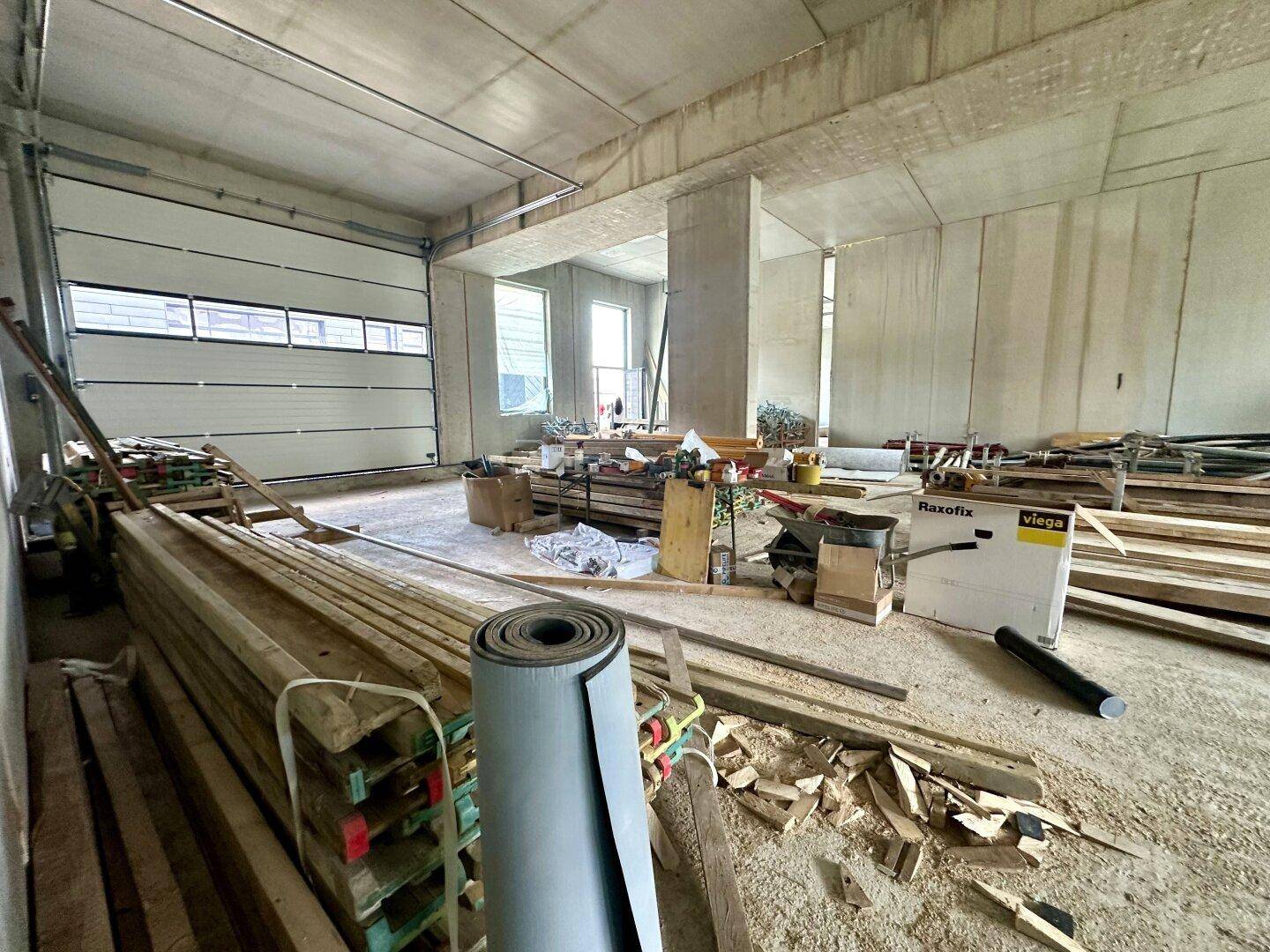 Geräumige Lagerhalle im Rohbau mit Baumaterialien, Rolltor und sichtbaren Betonwänden.