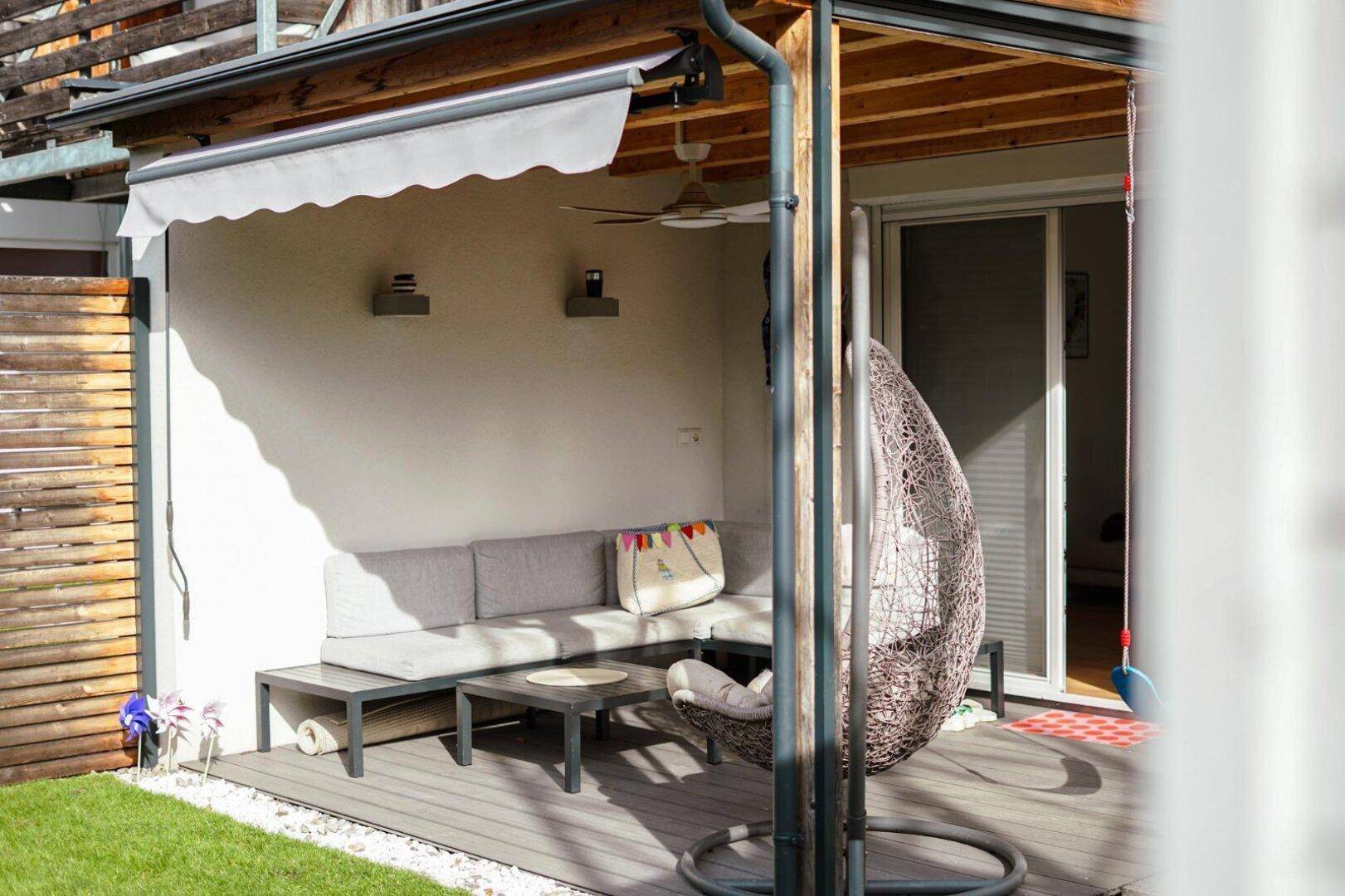 Gemütliche überdachte Terrasse mit Lounge-Möbeln und Hängesessel, angrenzend an einen kleinen Garten.