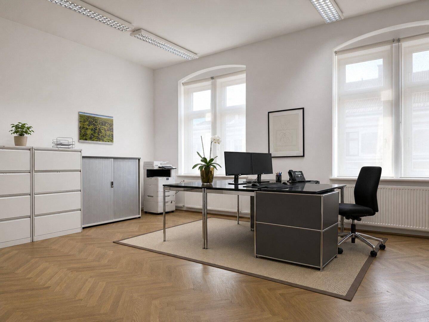 Modern eingerichtetes Büro mit hellem Fischgrätparkett, zwei großen Fenstern und funktionalen Büromöbeln.