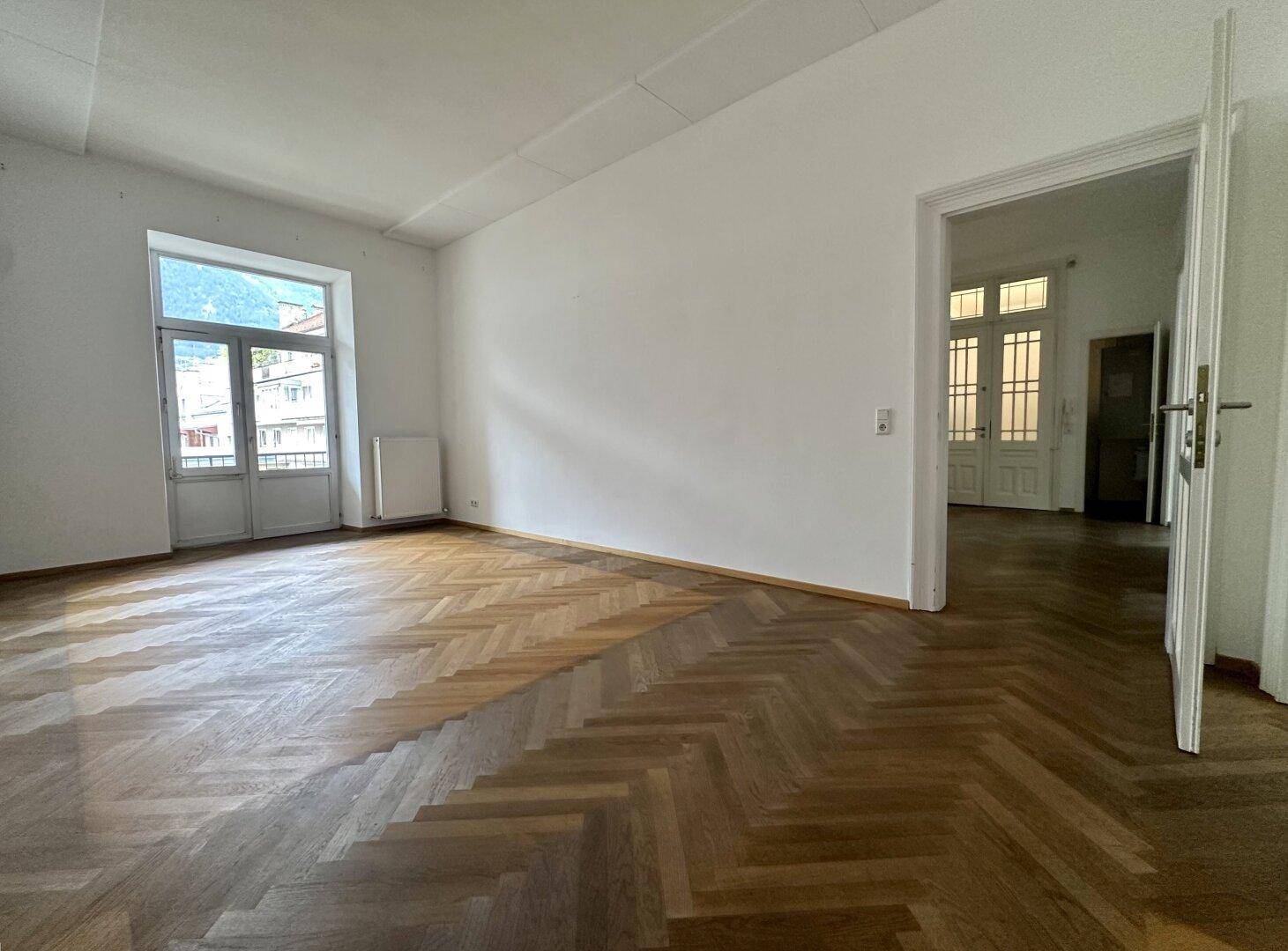 Heller Wohnbereich mit Fischgrätparkett und einer Doppeltür, die zu einem Balkon oder einer Terrasse führt.