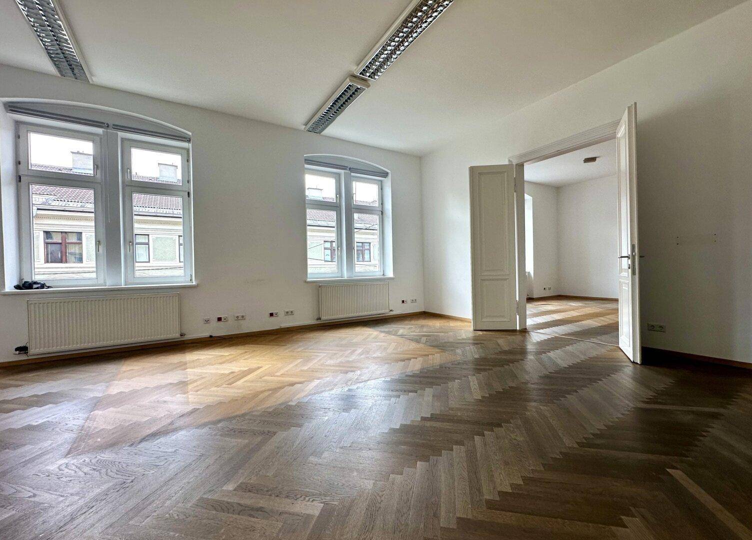 Geräumiger, leerer Raum mit großen Fenstern, die viel Tageslicht hereinlassen, und klassischem Fischgrätparkett.