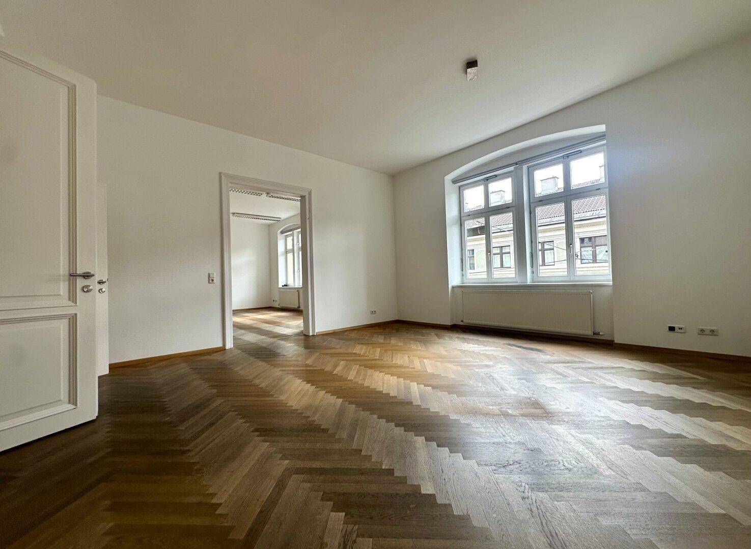 Heller Wohnraum mit klassischem Fischgrätparkett und hohen weißen Türen, die in einen Nebenraum führen.