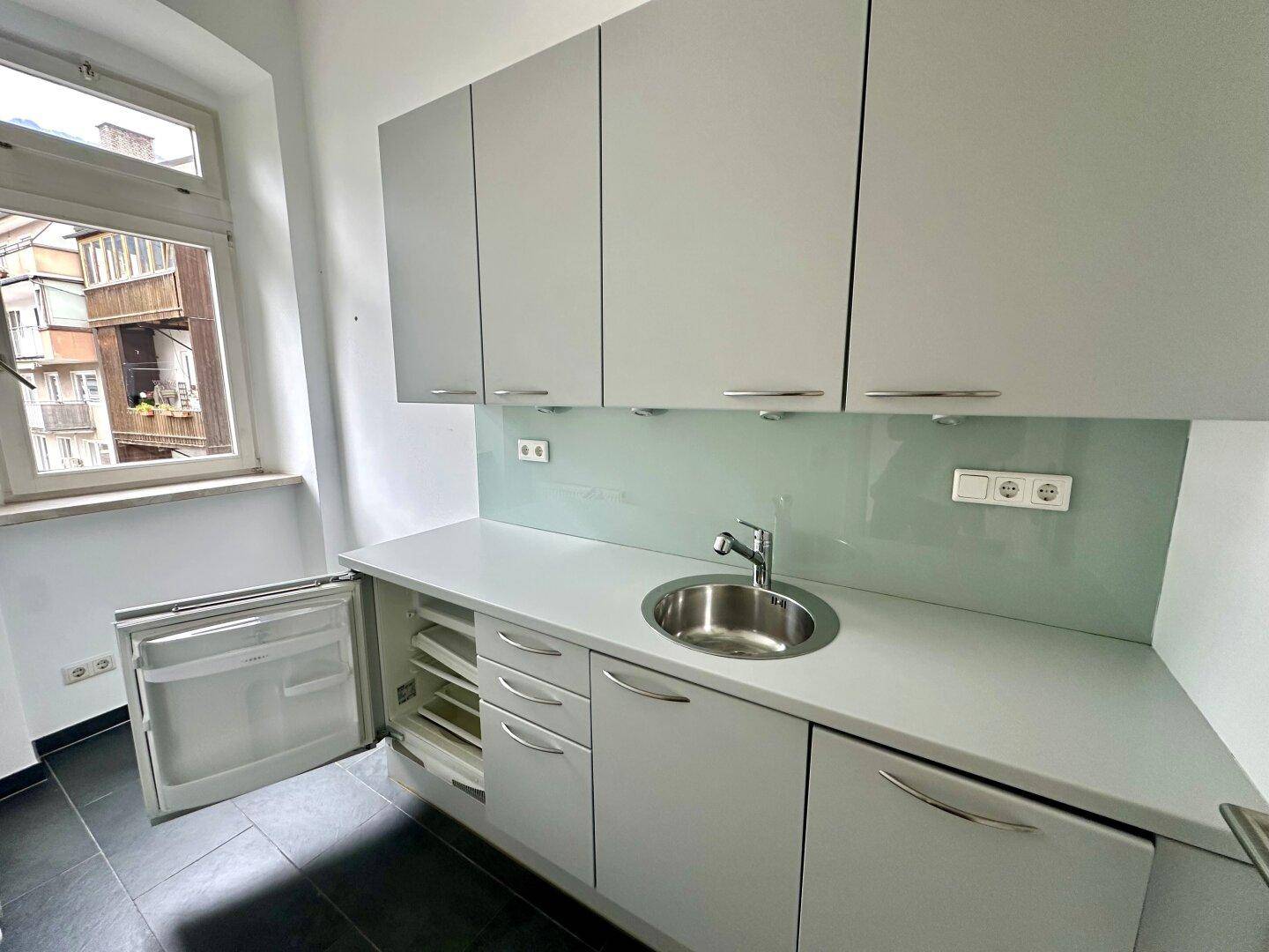 Funktionale Küchenzeile mit hellgrauen Schränken, Spüle und integriertem Kühlschrank unter dem Fenster.
