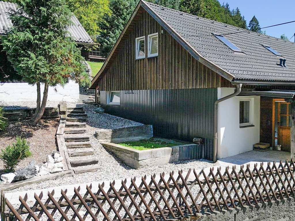 Seitenansicht eines Hauses mit Holzverkleidung, Satteldach und einem kleinen umzäunten Bereich.