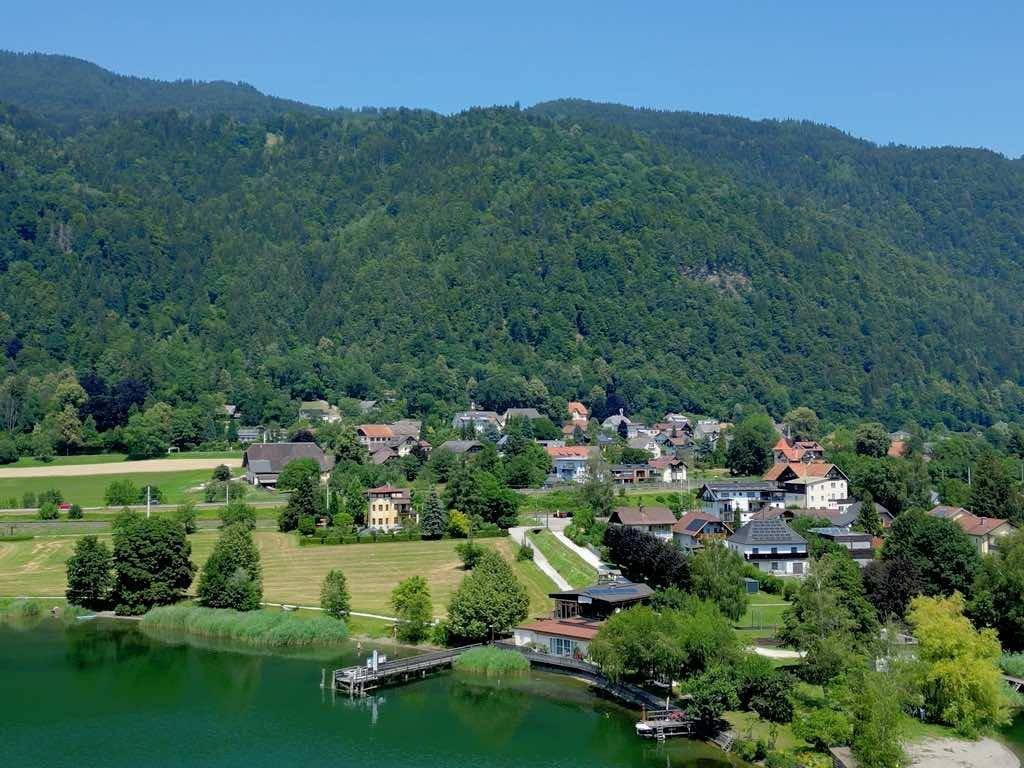 Panoramablick auf ein Dorf am Seeufer, eingebettet in eine grüne Berglandschaft.