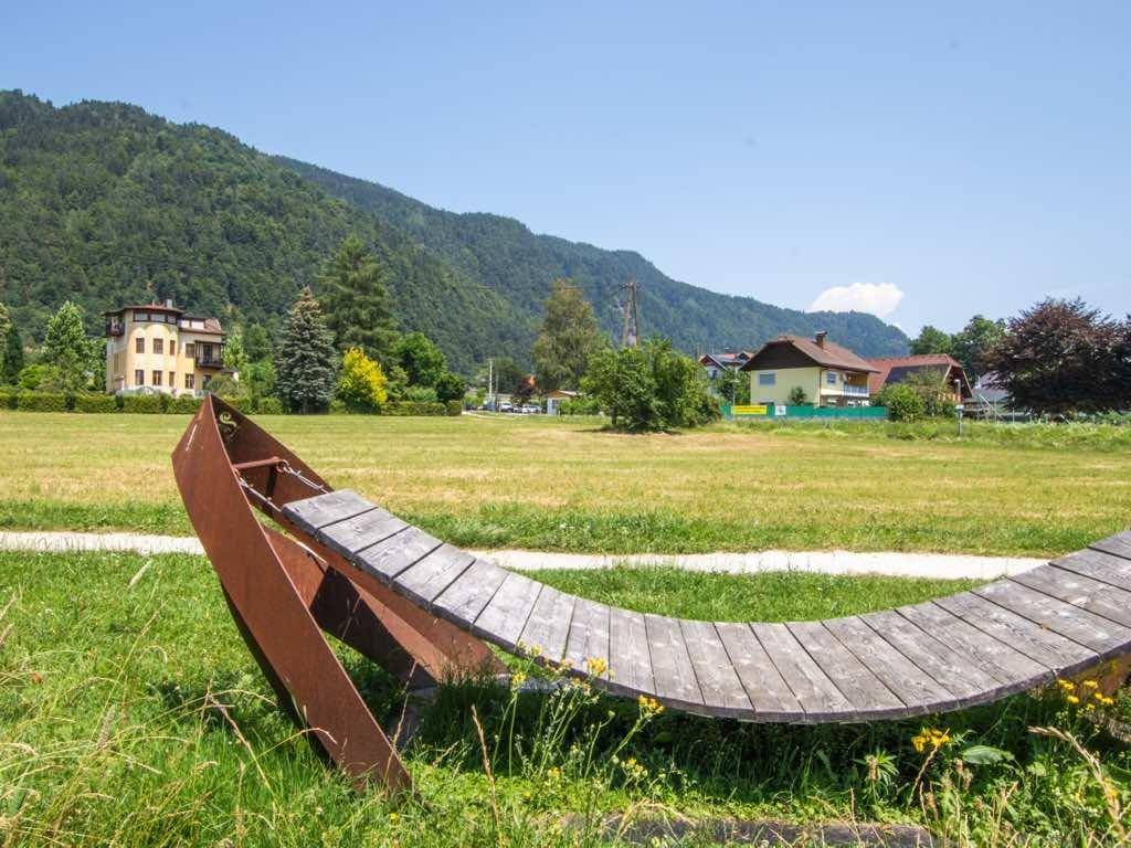 Geschwungene Holzbank in einer weiten Wiese, mit Häusern und bewaldeten Bergen im Hintergrund.