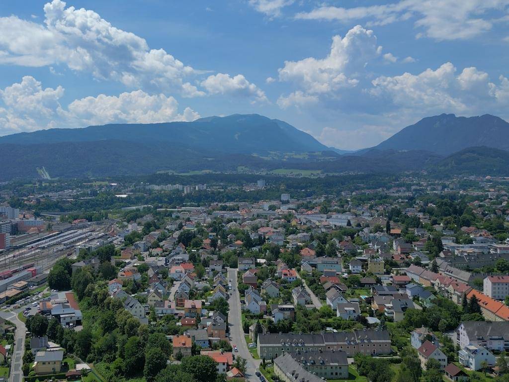 Panoramablick über eine Stadt mit grünen Hügeln und Bergen im Hintergrund unter blauem Himmel.