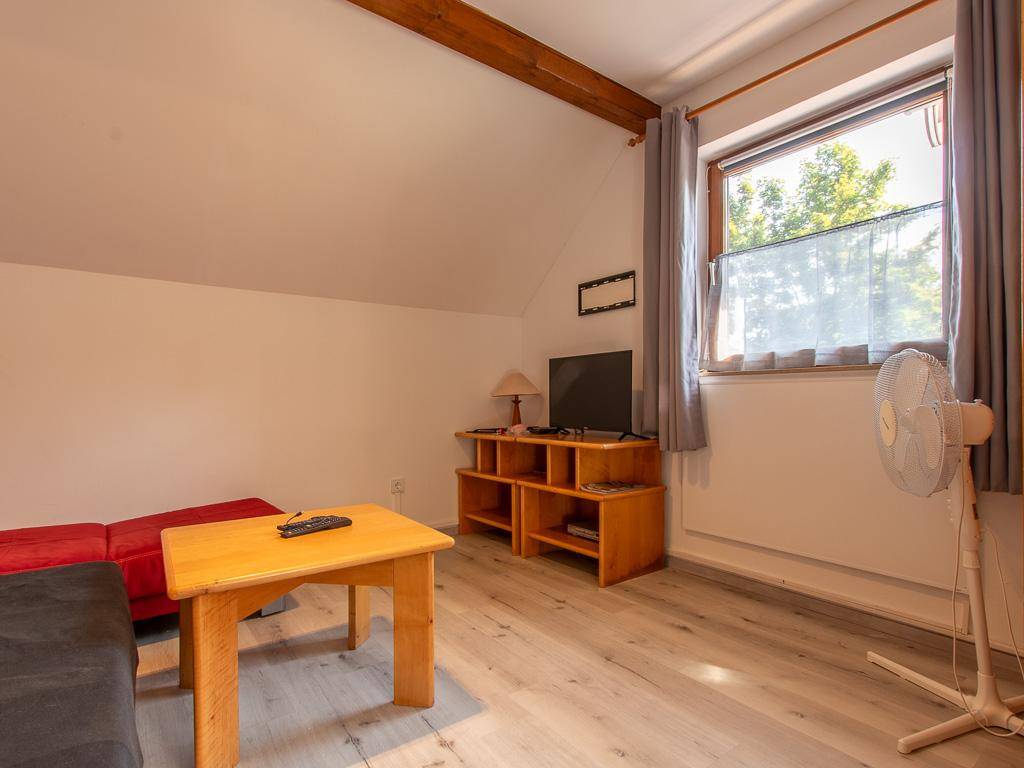 Einfaches Wohnzimmer im Dachgeschoss mit Sofa, Fernseher und Fenster mit Blick ins Grüne.