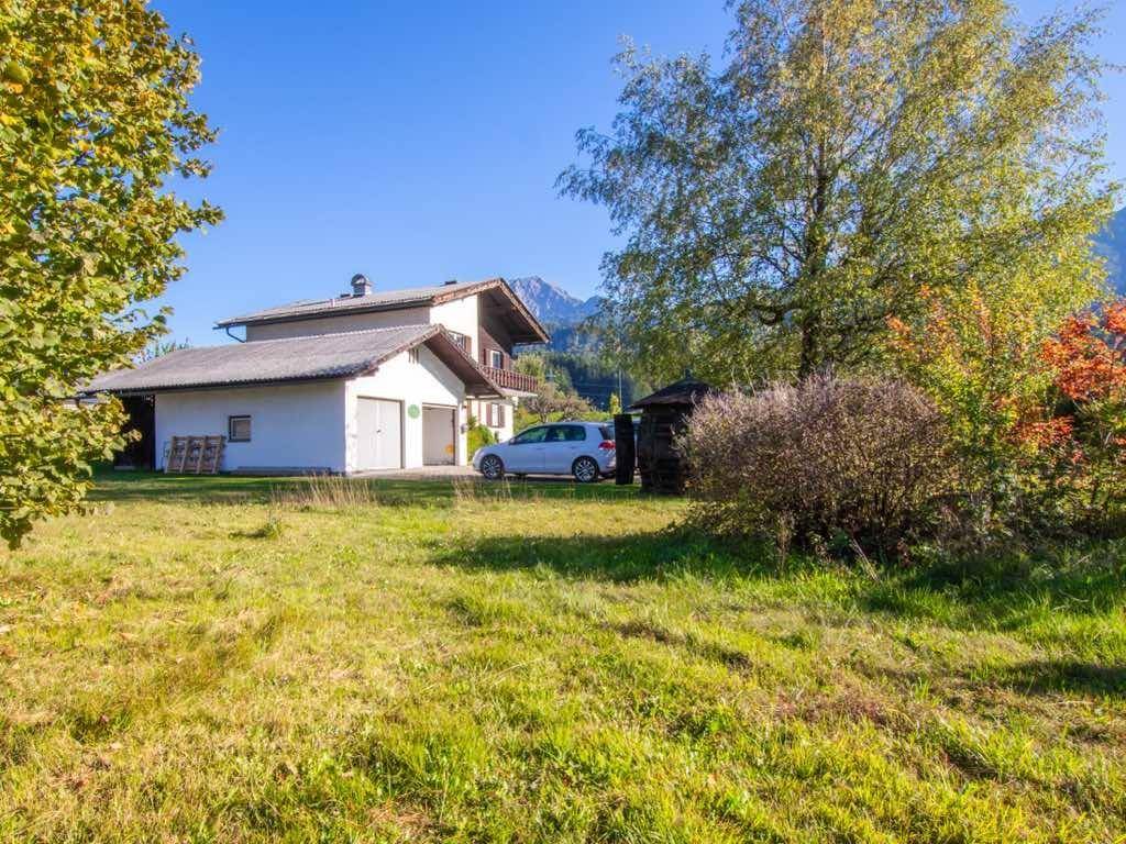 Hausfassade mit Doppelgarage und weitläufiger grüner Wiese vor einer beeindruckenden Bergkulisse.